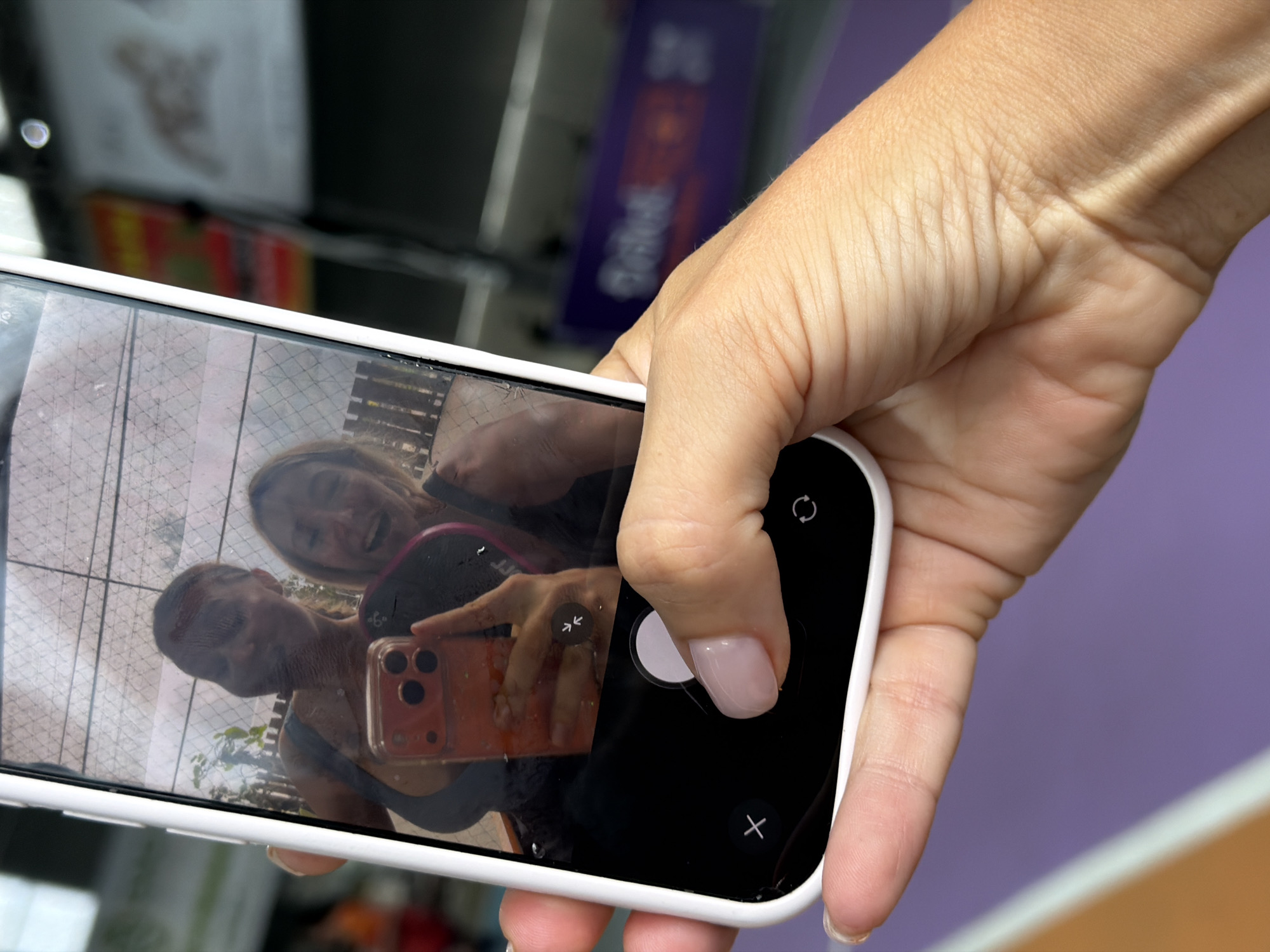 A person's hand, with a light pink manicured fingernail, is holding a white iPhone. The phone's screen displays a selfie of two women laughing, reflected in the screen. One woman has blonde hair and is in the foreground, while the other woman is in the background. The phone's camera app is open, showing a red-orange phone case. The background of the phone screen appears to be a fence and greenery. The image is taken from a low angle, showing the hand and phone against a blurred background with purple and orange hues. The overall scene appears to be outdoors in a public space, possibly a park or recreational area in Da Lat, Vietnam.