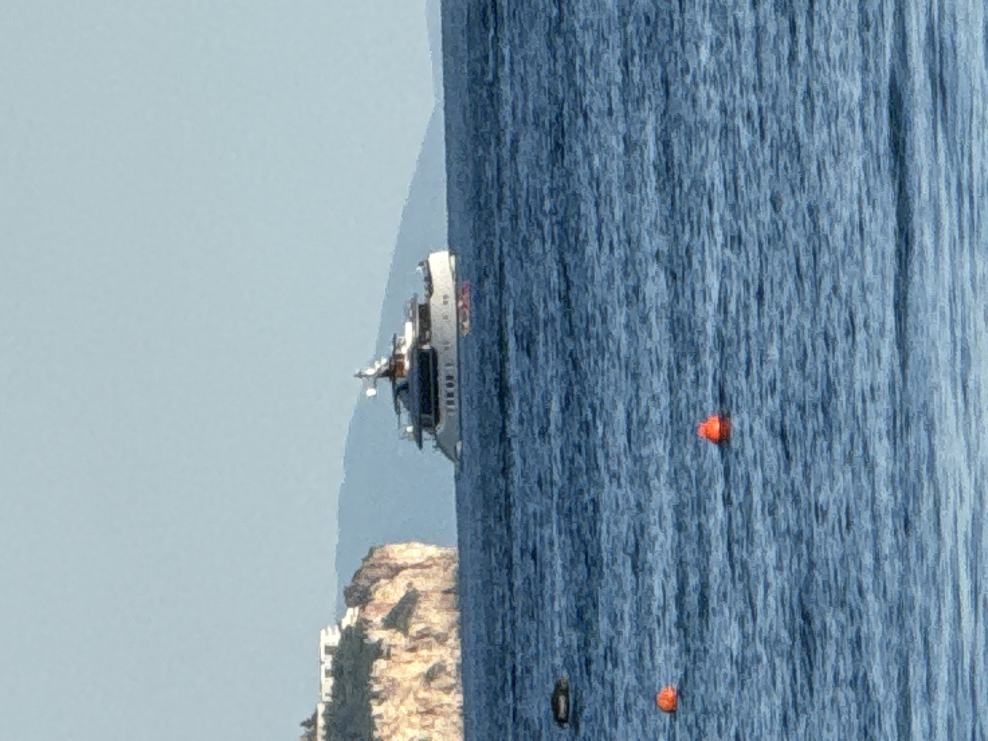 A wide shot shows a large white yacht on the blue ocean. Behind the yacht, a tan cliff with buildings on top is visible, along with hazy mountains. In the foreground, two orange buoys float on the water. The sky is a clear light blue.