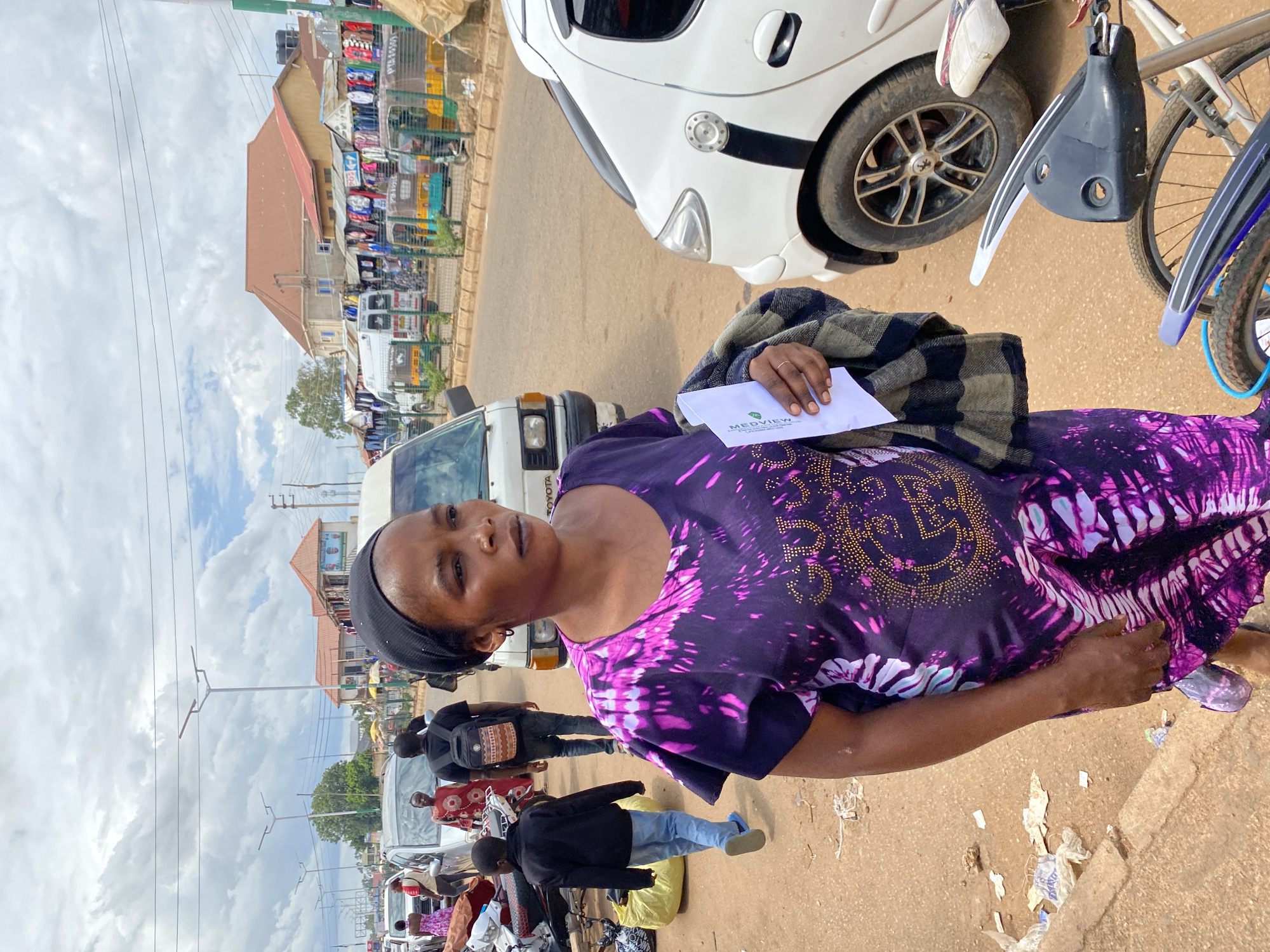 A medium shot captures a Black woman in a bustling outdoor marketplace in Bukuru, Nigeria. She is positioned in the foreground, facing slightly to her right, with her head turned to look towards something outside the frame. She wears a black headscarf and a vibrant purple and pink tie-dye dress adorned with gold embellishments. In her right hand, she clutches a white paper document and a folded grey and black checkered blanket.
Behind her, the marketplace teems with activity. Several vehicles are visible, including a white minivan and a white car. A young Black boy in a black jacket and blue jeans walks away from the camera in the midground. Other individuals, including women in traditional attire and a man with a backpack, are scattered throughout the scene. Stalls with various goods, including clothing displayed on racks, line the street, and an assortment of motorcycles and tricycles are parked or in motion. The ground is a dusty, reddish-brown earth, littered with some debris. The sky above is overcast with dramatic cloud formations. The overall impression is one of a lively, everyday scene in a Nigerian urban environment.