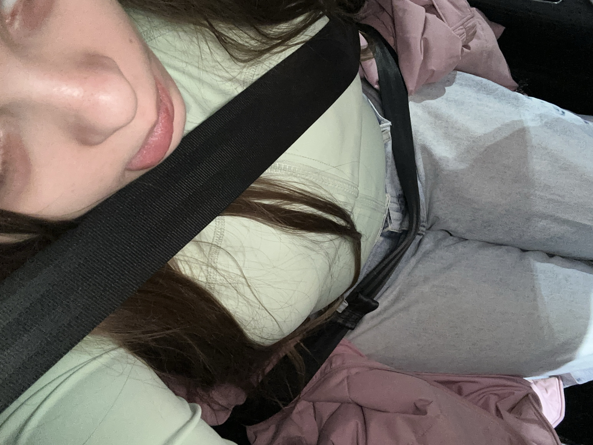 A close-up, slightly angled shot shows a person wearing a seatbelt in what appears to be a car. The person has fair skin, brown hair, and is wearing a light green top and light blue jeans. Part of their face, including their nose and lips, is visible. A black seatbelt diagonally crosses their body. A pink jacket is partially visible below the seatbelt and jeans. The background is dark and indistinct. There is no information in the image to determine the location in Moscow, Russia.