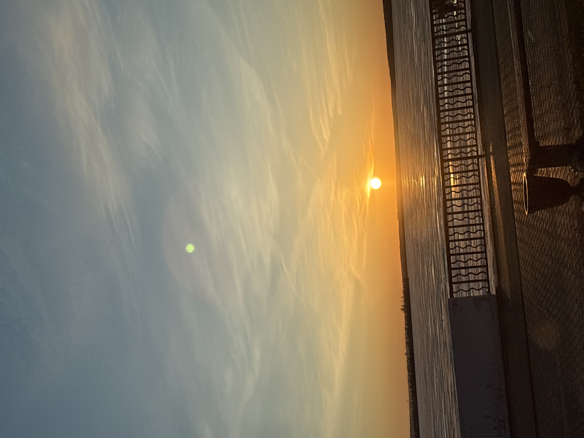 The image is presented rotated 90 degrees counter-clockwise from its natural landscape orientation. When viewed correctly, it captures a serene sunset or sunrise scene over a body of water in Votkinsk, Russia, with an adjacent embankment.
The sky dominates the upper portion, transitioning from a soft blue on the left to a brilliant orange and golden hue around the sun, which is low on the horizon. Wispy, delicate white clouds stretch across the sky, catching the warm light. A distinct, bright green circular lens flare is visible in the upper-left quadrant of the sky.
The sun, a vivid orange-yellow orb, casts a shimmering path of light across the calm body of water, likely a reservoir or river, which occupies the central and right-hand side of the main composition. Along the right edge of the water, a dark, ornate metal railing with a distinctive wavy pattern forms a barrier. Beyond the railing lies a paved walkway or embankment constructed from rectangular blocks. Further to the right on this paved area, a dark, silhouetted structure, possibly a lamp post or waste bin, is partially visible.
The scene depicts the peaceful "golden hour" during either sunrise or sunset, characterized by the warm, intense light and reflections. No people or specific activities are visible. There is no visible text in the image.