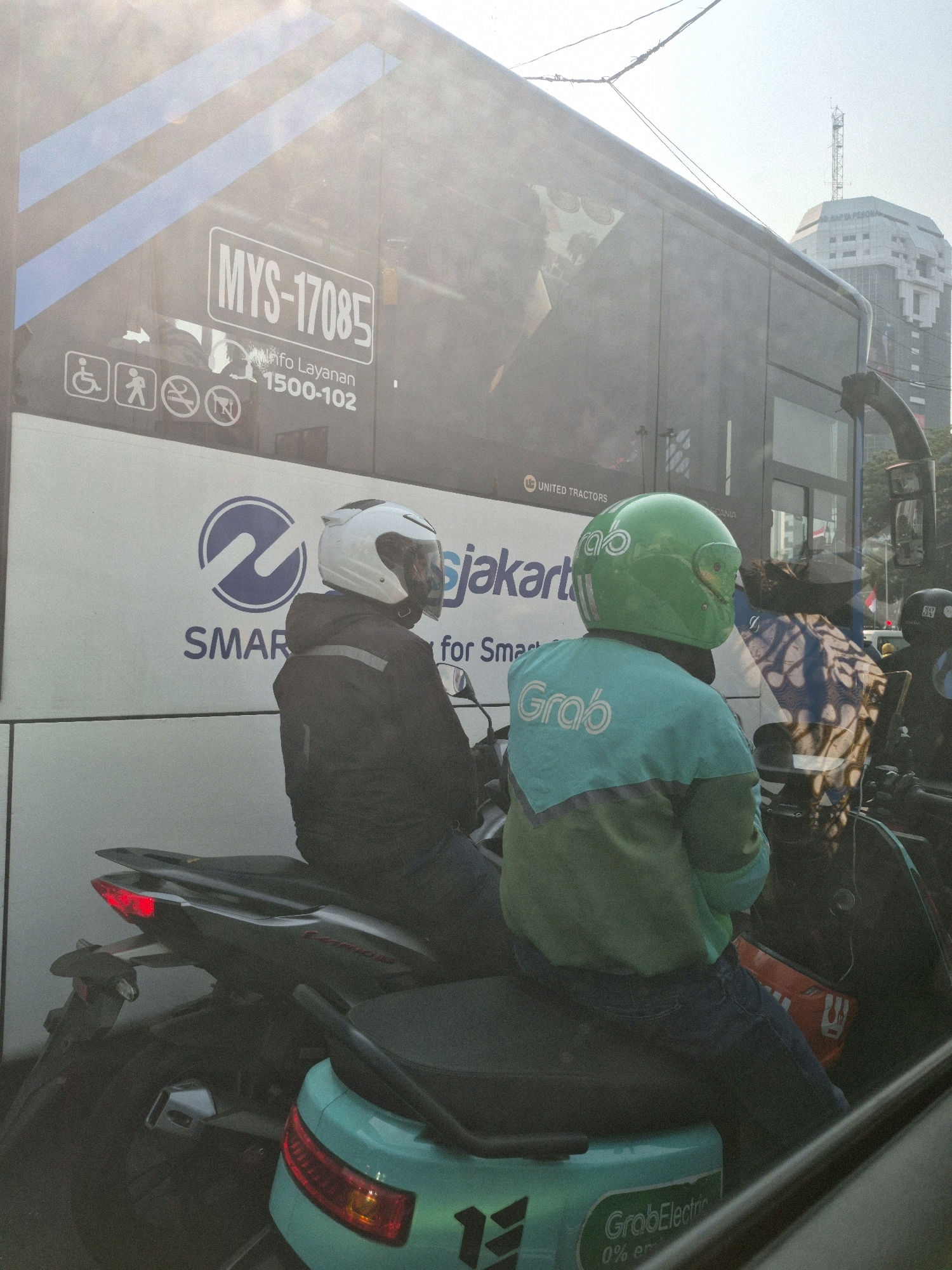 The image depicts a traffic scene in Central Jakarta, Indonesia, with two motorcyclists visible in the foreground, partially obscured by a bus. Both riders are wearing helmets. The rider on the left wears a dark jacket, and the rider on the right wears a teal jacket with "Grab" printed on the back. The bus behind them features various decals and text, including a license plate "MYS-17085" and logos related to "SMART CITY JAKARTA" and "UNITED TRACTORS." The background shows a hazy cityscape with buildings and power lines, suggesting an urban environment. The lighting indicates it is daytime, possibly during a congested period given the presence of multiple vehicles. The overall scene suggests a typical urban commuting or delivery context in Jakarta.