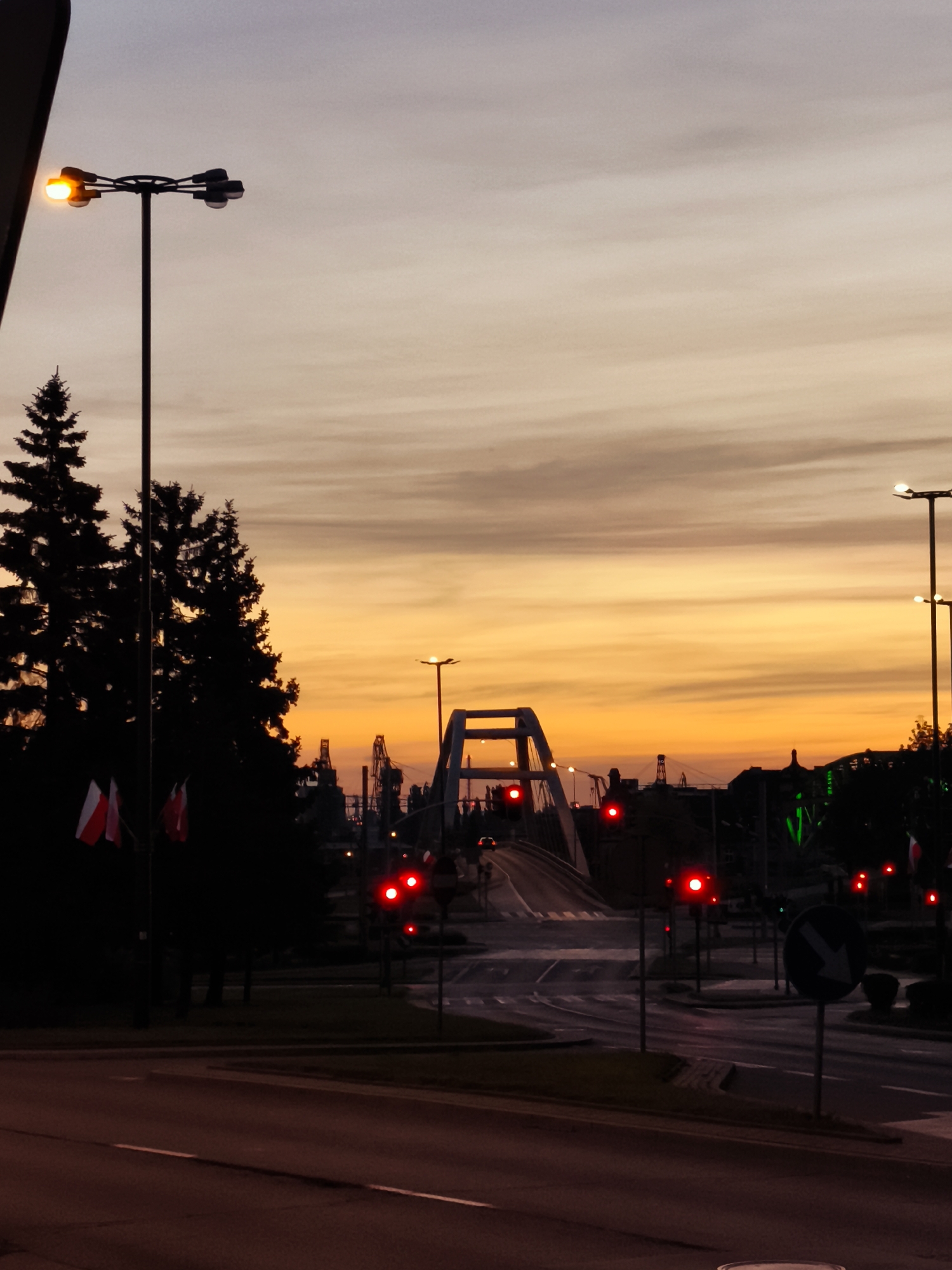 The image depicts an urban scene at dusk in Gdańsk, Poland, with a dramatic sky painted in hues of orange, yellow, and purple. A prominent feature is a modern bridge arching over a road, illuminated by the fading light and streetlights. Several traffic lights are visible, with some displaying red signals, indicating stopped traffic. Silhouetted against the sky are tall evergreen trees and what appear to be industrial cranes in the distance, suggesting the proximity of a port or industrial area. Two flags, likely the Polish flag, are visible on poles near the trees. The foreground shows a road with lane markings. The overall atmosphere is one of transition from day to night, with a sense of urban activity and infrastructure.