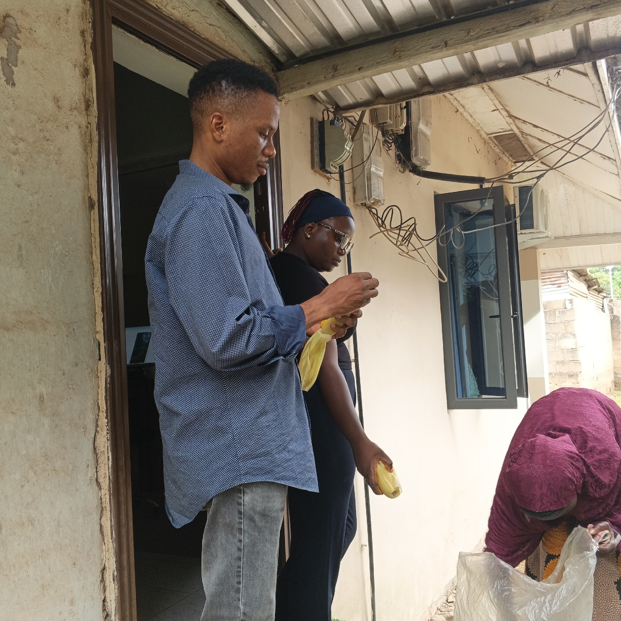 In Shaba, Nigeria, a group of individuals stands near an open doorway. A man in a blue dotted shirt and grey jeans holds a yellow object. To his right, a woman wearing a dark top and a blue headscarf is looking at her hands, also holding a yellow object. In the foreground, a person in a purple head covering and patterned skirt is bending over a transparent plastic bag. The scene is set against a building with a corrugated metal roof and visible electrical wiring and meters.