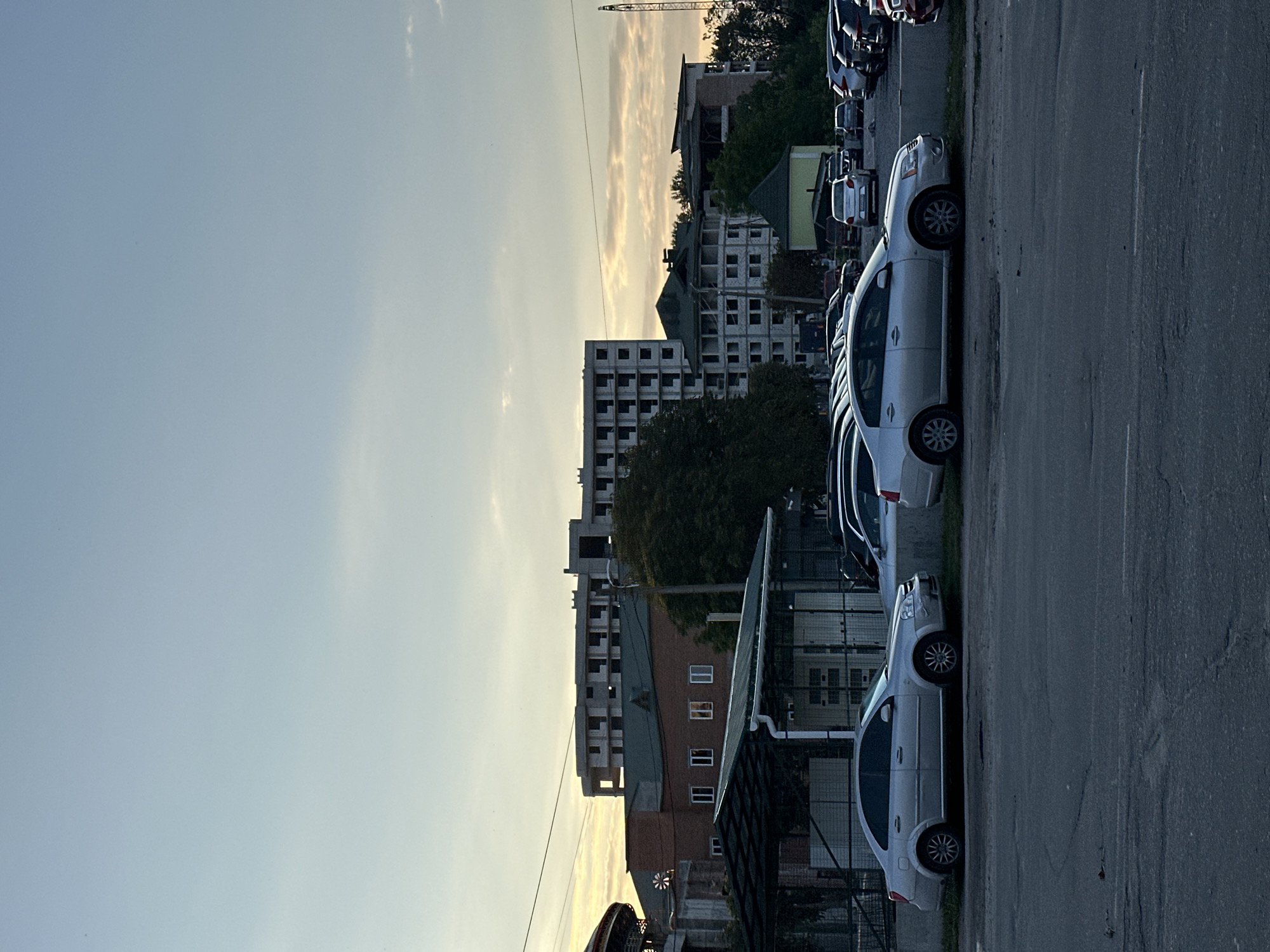 In the city of Khmilnyk, Ukraine, a parking lot is visible with several cars, including a silver sedan in the foreground. Behind the parking lot, a two-story building with a green roof and a covered area with a green roof are present. Further in the background, several unfinished multi-story buildings under construction are observable. The sky is light blue with some clouds and the sun is setting, casting a warm glow. A construction crane is visible in the far right of the image.