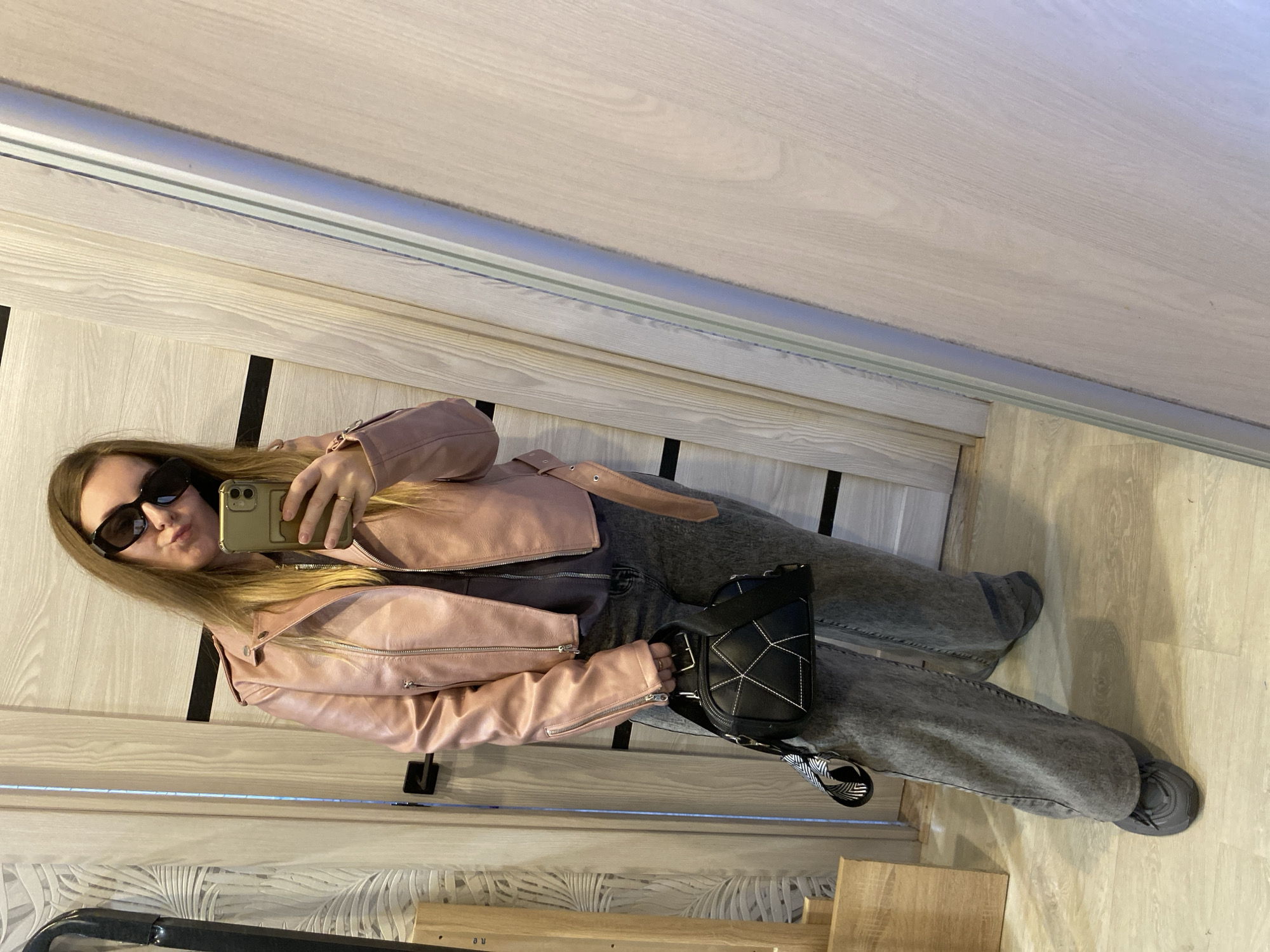 A young woman with long blonde hair and sunglasses is taking a selfie in a mirror. She is wearing a pink leather jacket, gray top, and gray jeans with sneakers. She is holding a gold-colored smartphone in her right hand and a black bag with white stitching is slung across her body. The reflection shows a light-colored wooden surface with darker vertical stripes, possibly a door or paneling. The location is Noviy, Russia.