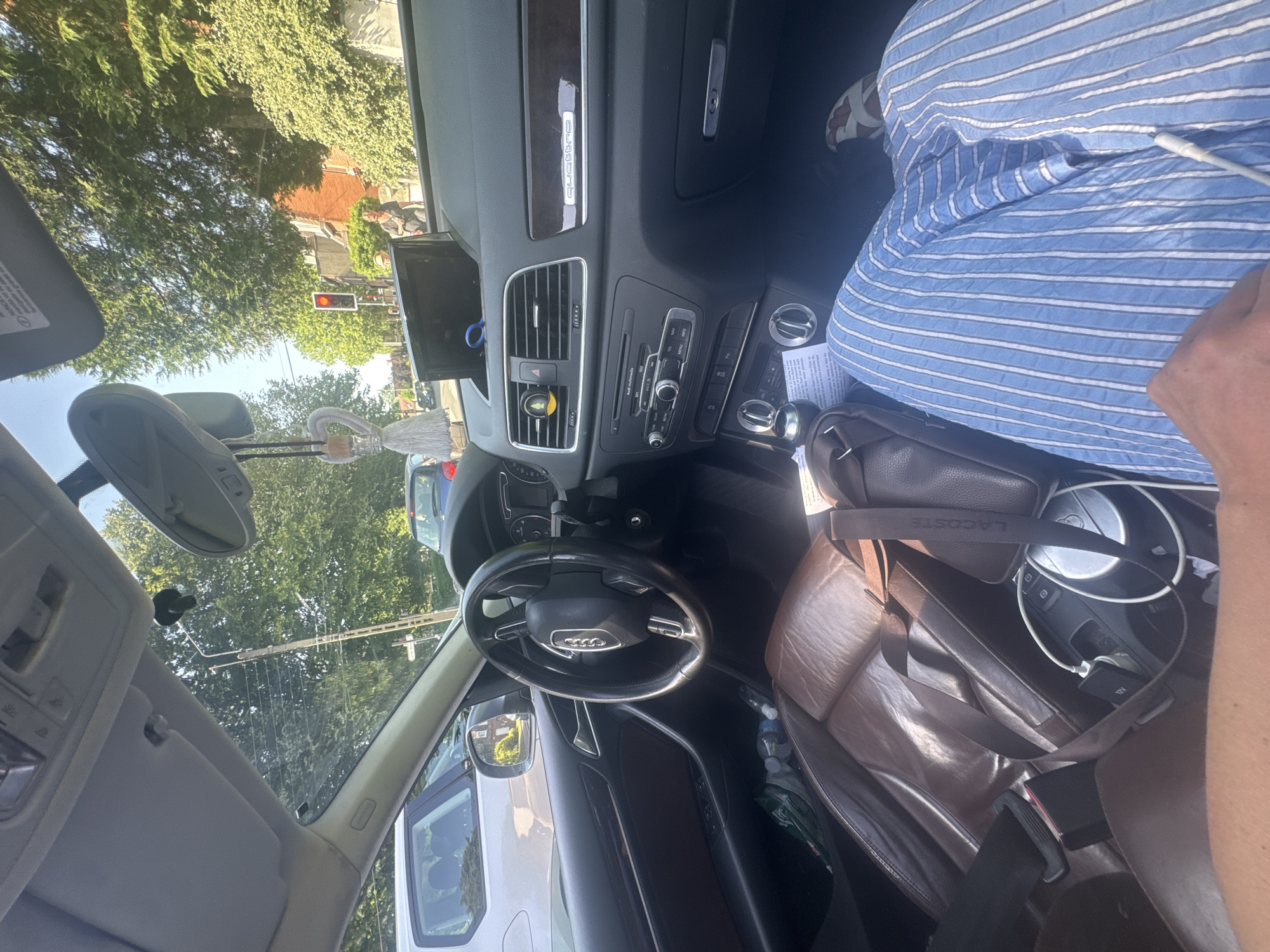 The image shows the interior of an Audi Q3, likely taken from the driver's seat. The vehicle is stopped at a red traffic light, as seen through the windshield. In front, there is a white delivery van with reflections of trees visible on its windows. The Audi's dashboard is in view, showing the infotainment screen and climate control panel. A Lacoste leather bag sits on the passenger seat, obscuring part of the blue and white striped shirt worn by the passenger. A dangling tassel-style ornament hangs from the rearview mirror. The outside environment is characterized by green trees and residential buildings, typical of a suburban neighborhood. The location is likely Marcq-en-Barœul, France, based on contextual information provided.