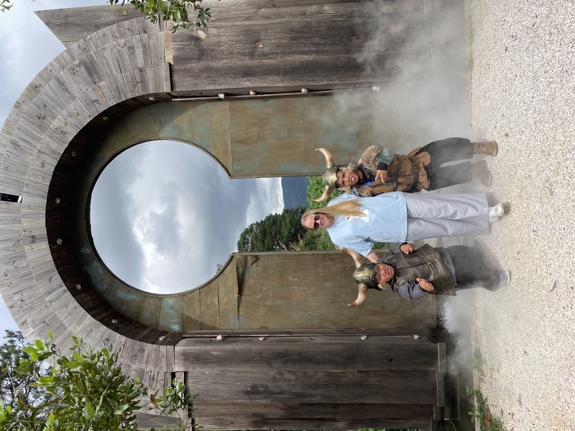 A woman is standing between two men dressed as Vikings in front of a large, wooden archway with a circular opening. The archway is made of weathered wood and has metal accents. The two men are wearing horned helmets and Viking-style armor. One man is giving a thumbs-up gesture. Smoke or fog is present around the feet of the men. The ground is covered in white gravel. In the background, there are trees and a cloudy sky. The image appears to be taken outdoors in a park or themed attraction, potentially in the city of Thôn 1, Vietnam, given the context provided.