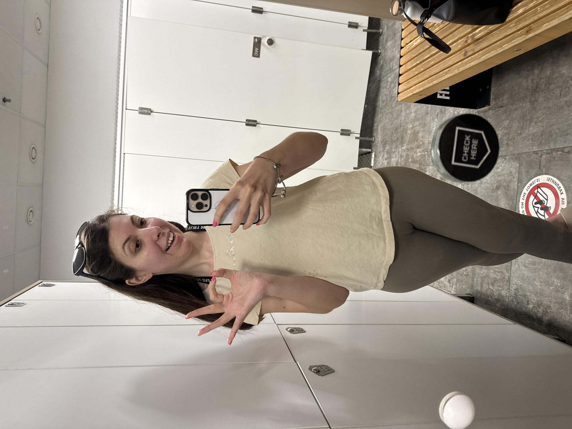 A woman is taking a selfie in a locker room. She is wearing a light tan t-shirt and grey leggings. She has dark brown hair with sunglasses perched on top and is making an "okay" gesture with her left hand. She is holding her phone in her right hand, which is partially visible. In the background, there are white lockers and stalls. A wooden bench with a black bag and water bottle on it is visible on the right side of the image. There is a circular sticker on the floor that says "CHECK HERE". The lighting is bright, and the overall environment appears to be a gym or fitness facility.