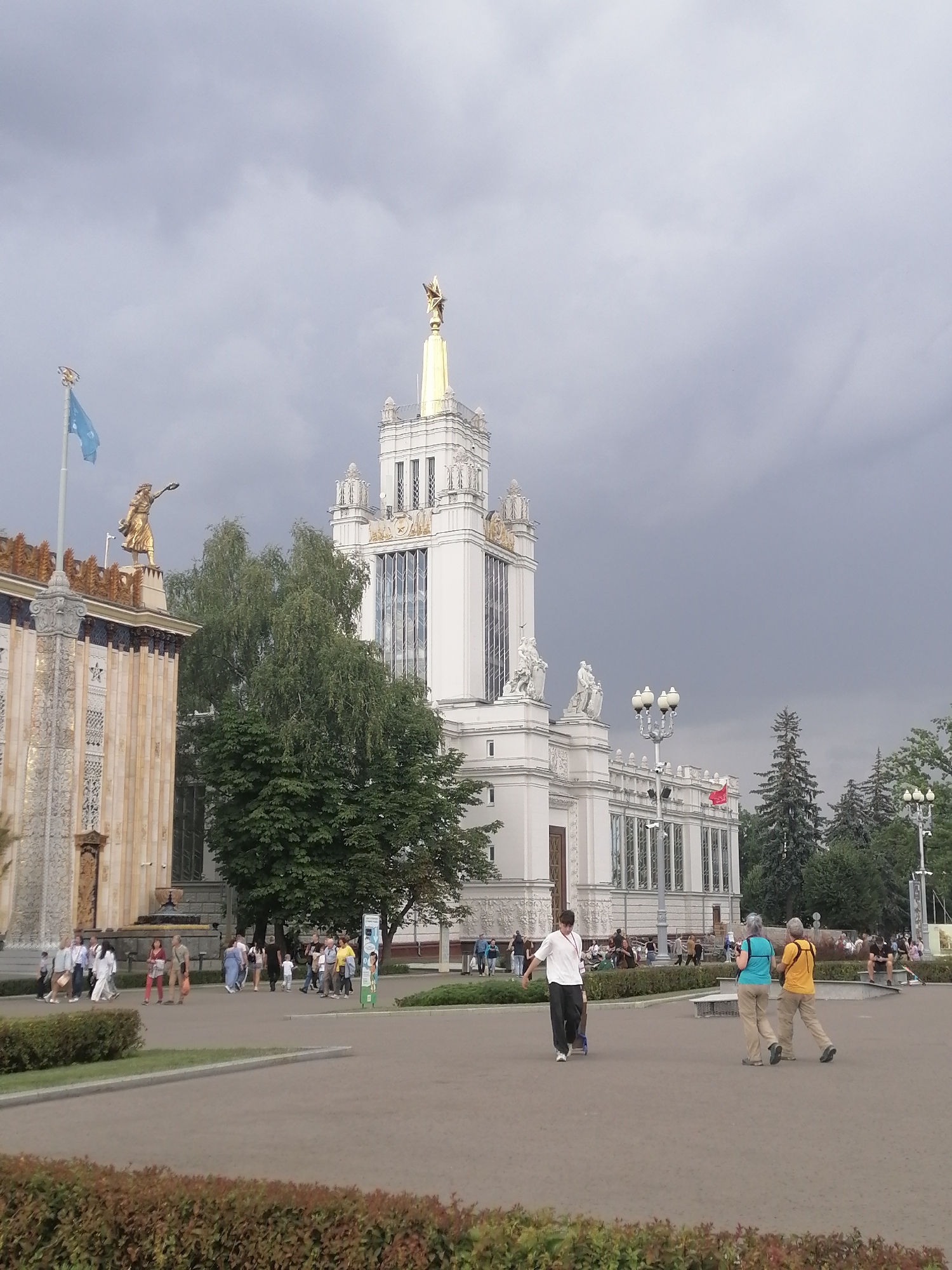 The image captures an outdoor scene at VDNKh (Exhibition of Achievements of National Economy) in Moscow, Russia, under a cloudy sky. In the background, a grand, white, Soviet-era pavilion with ornate decorations and a tall, golden spire stands prominently. To the left, another pavilion with intricate detailing and a golden statue on top is visible, along with a blue flag on a pole. In the mid-ground, a group of people are walking around the paved area. In the foreground, a young person is pulling a blue cart, while two women are walking away from the camera. Lush green trees and bushes are scattered throughout the scene, adding to the park-like atmosphere of the exhibition grounds.