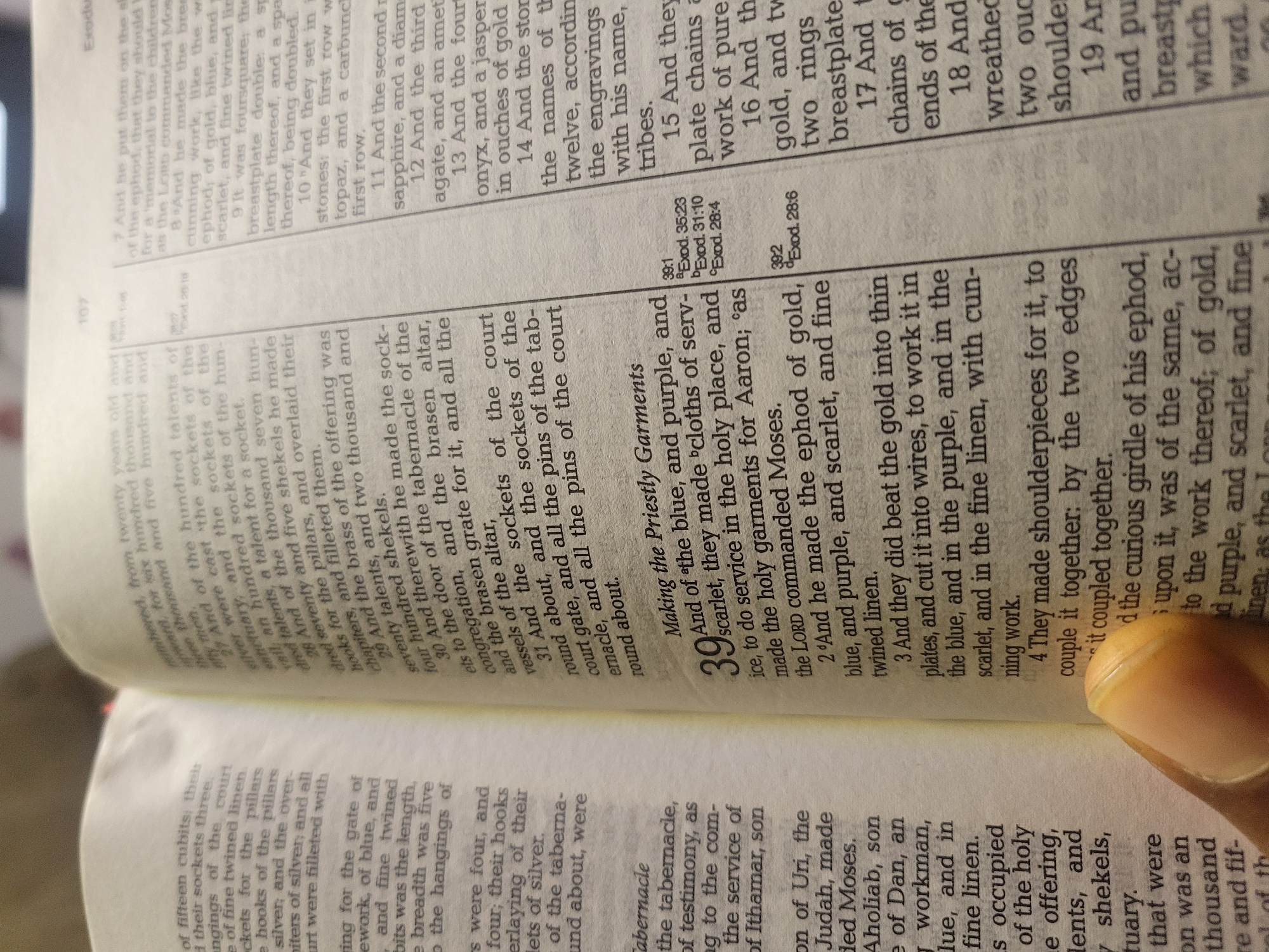 This image shows a close-up view of pages from a book, likely a Bible. The pages display printed text in a serif typeface, organized into paragraphs and numbered verses. Chapter 39 of the book of Exodus is visible, detailing the making of priestly garments. A hand and fingertips are visible in the lower right corner of the image. The book appears to be open on a flat surface, with the pages slightly curved at the spine. The photograph appears to have been taken in indoor lighting, given the shadows and contrasts in the image.