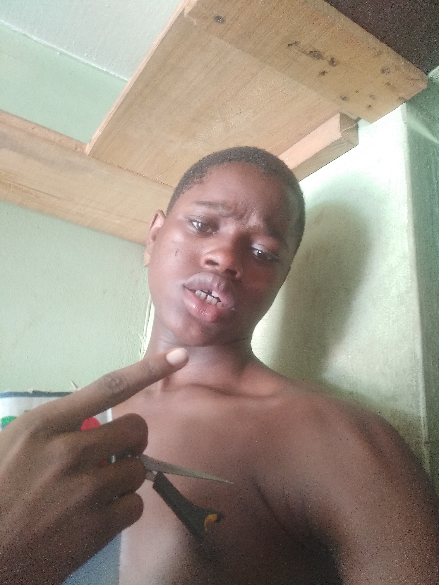 A dark-skinned person with short hair holds a pair of scissors in their left hand. The person is shirtless and stands indoors, with a wooden structure above them and a greenish wall in the background. The location is Shangisha, Nigeria.