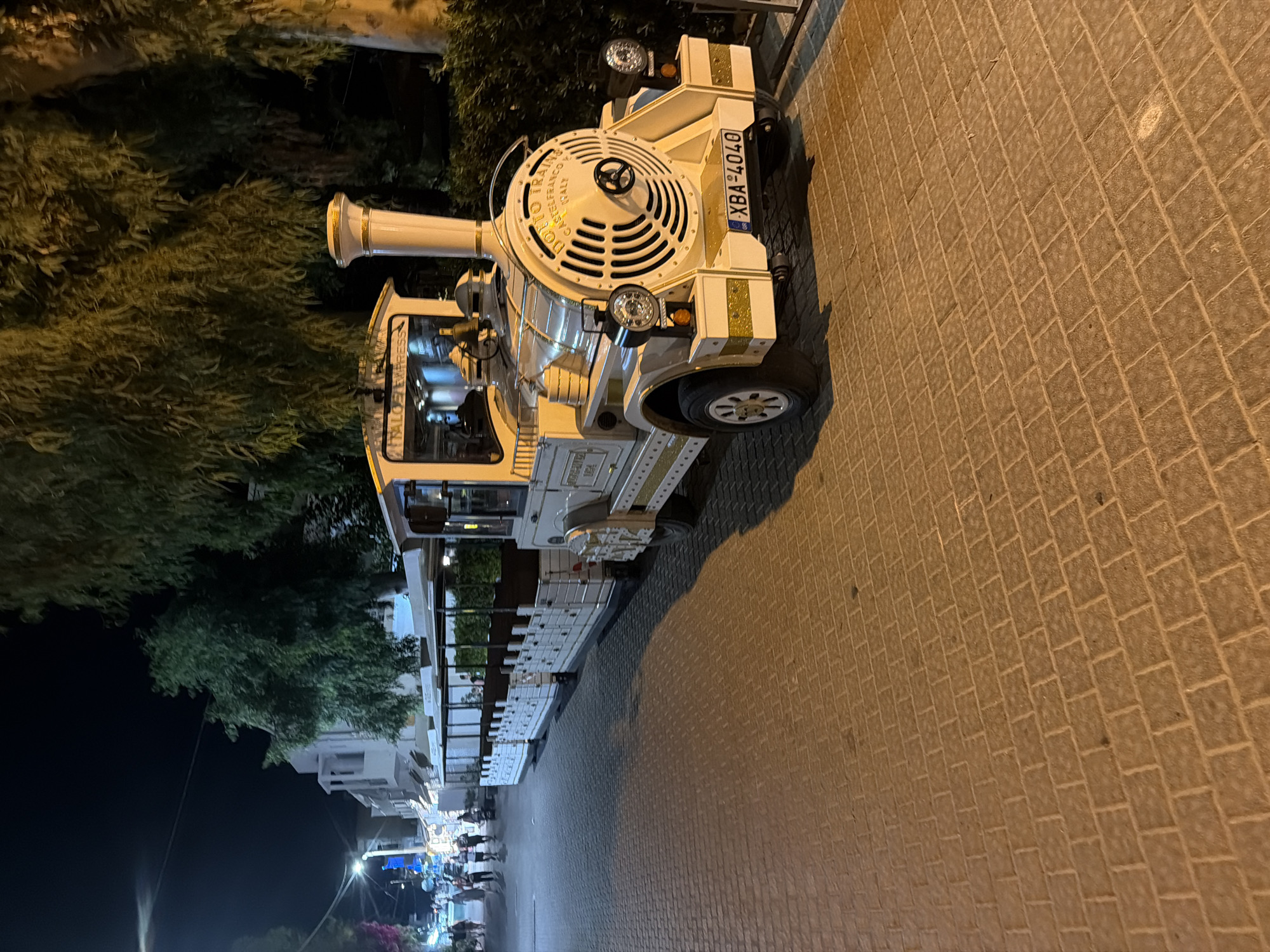 In Georgioupoli, Greece, a white tourist train, identified as "DOTTO TRAINS CASTELFRANCO ITALY," is parked on a cobblestone street at night. The train consists of a locomotive and several passenger carriages, all adorned with gold accents. The locomotive features a circular grille with the brand name and "ITALY" visible, along with headlights and a license plate reading "GR XBA-4040." Adjacent to the train, several people are walking on the sidewalk, and shops with awnings line the street. Lush green trees and foliage are present in the background, and streetlights illuminate the scene, casting shadows on the ground. The overall environment suggests a lively tourist area during evening hours.