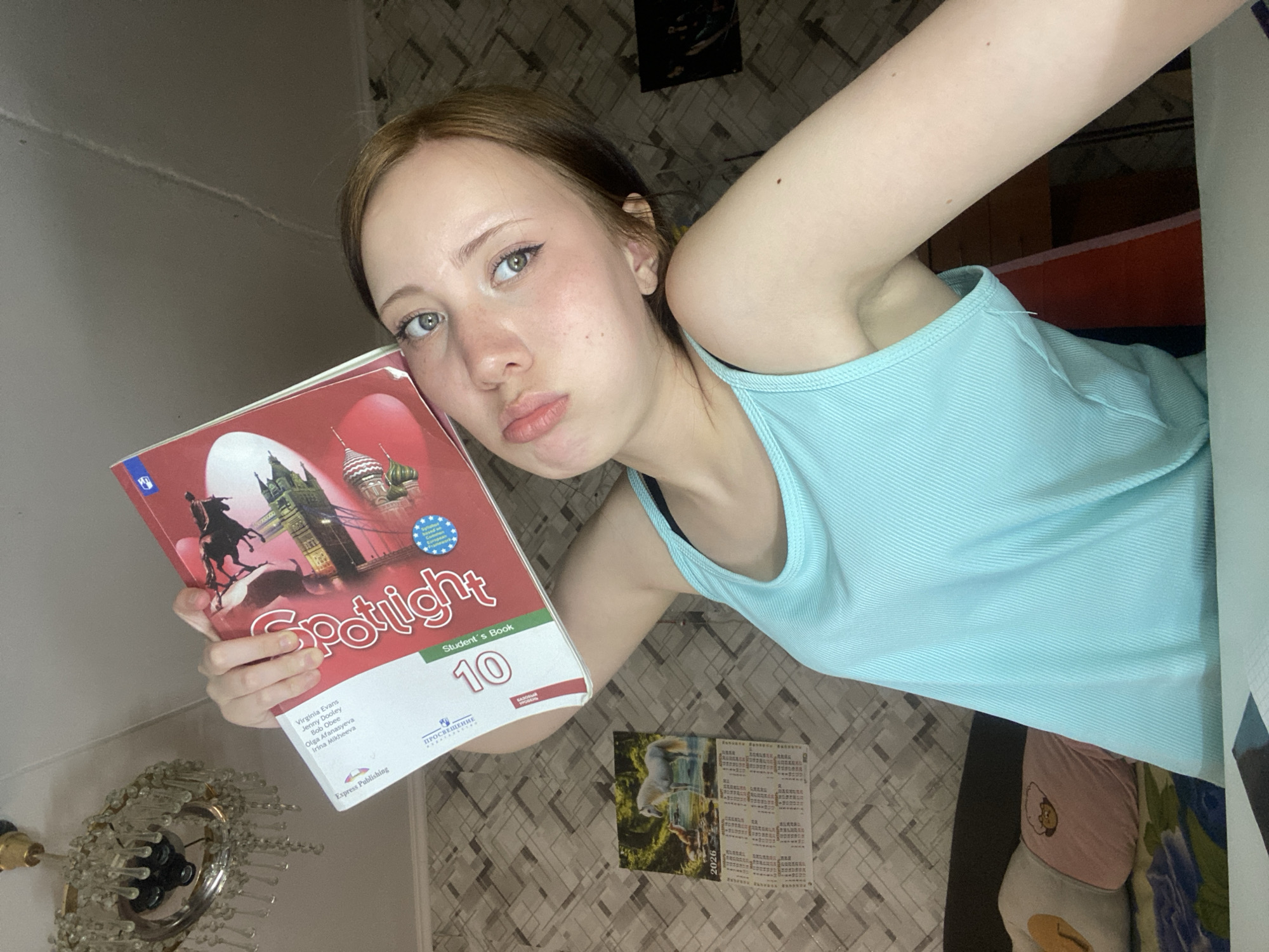A young woman is holding a textbook titled "Spotlight 10" and has a somewhat pouty expression on her face. She is wearing a light blue tank top. The background appears to be a room with textured walls, a calendar, and a decorative light fixture. The image is taken from a slightly high angle.