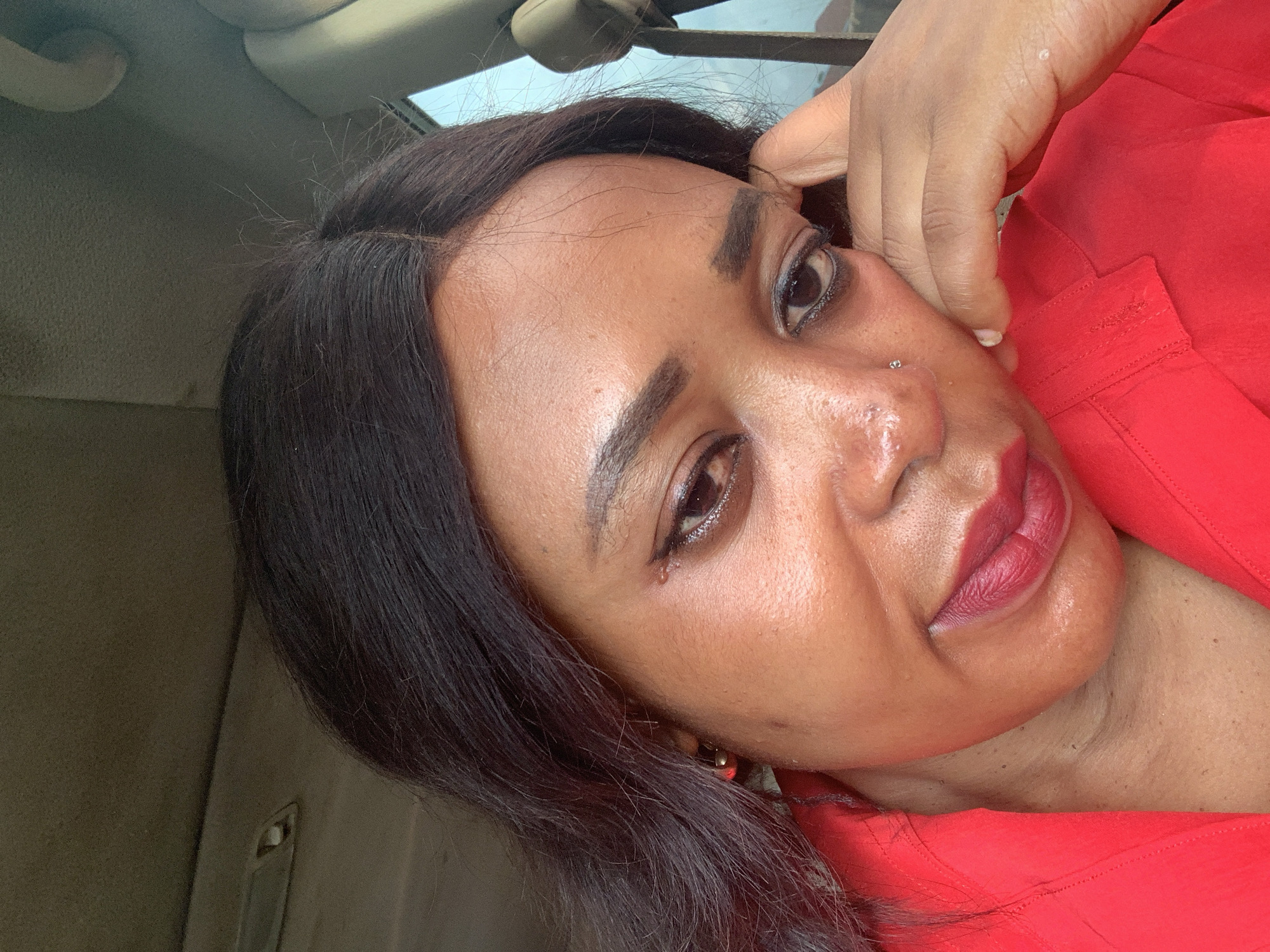 A close-up, outdoor selfie shows a Black woman in a red collared shirt, resting her right cheek on her hand. Her dark hair is parted on the left and cascades over her left shoulder. She wears dark eyeliner and red lipstick. A small nose ring is visible. The background suggests the interior of a vehicle, with the ceiling and a portion of the door visible. The scene is presumed to be in Sabon Tasha, Nigeria.