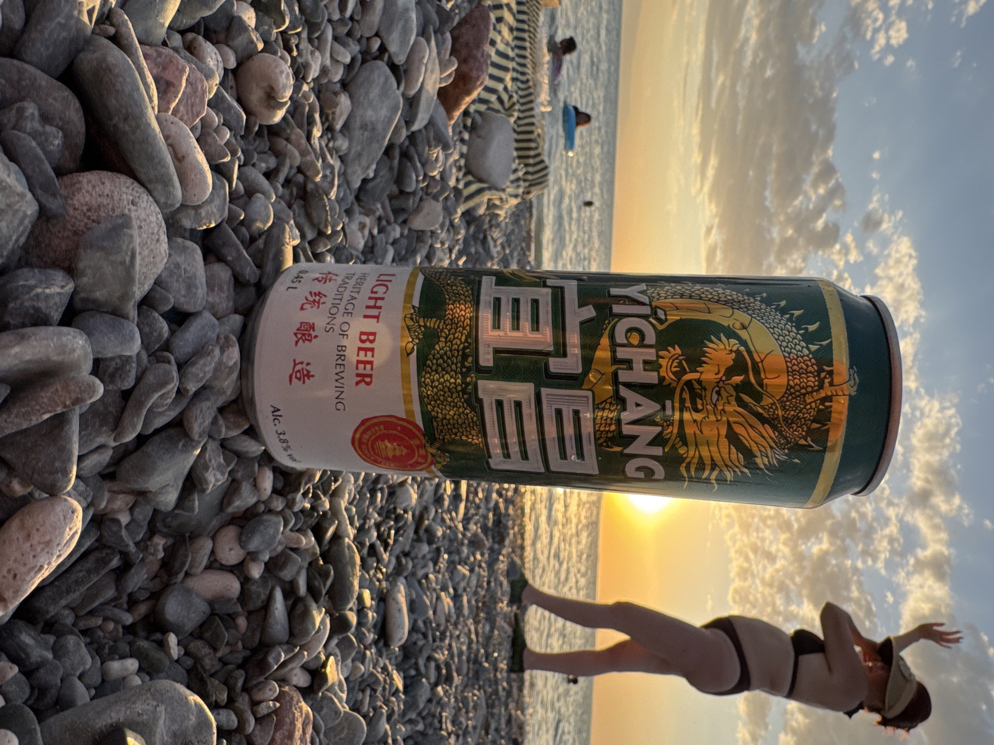 A can of Yichang Light Beer stands upright on a pebbled beach in Zharbulak, Kazakhstan, with the Caspian Sea in the background under a cloudy sunset sky. A woman in a bikini and sun visor is silhouetted against the setting sun, her arm raised. Two individuals are visible in the water, one with a blue float. The foreground consists of numerous grey and pinkish pebbles.