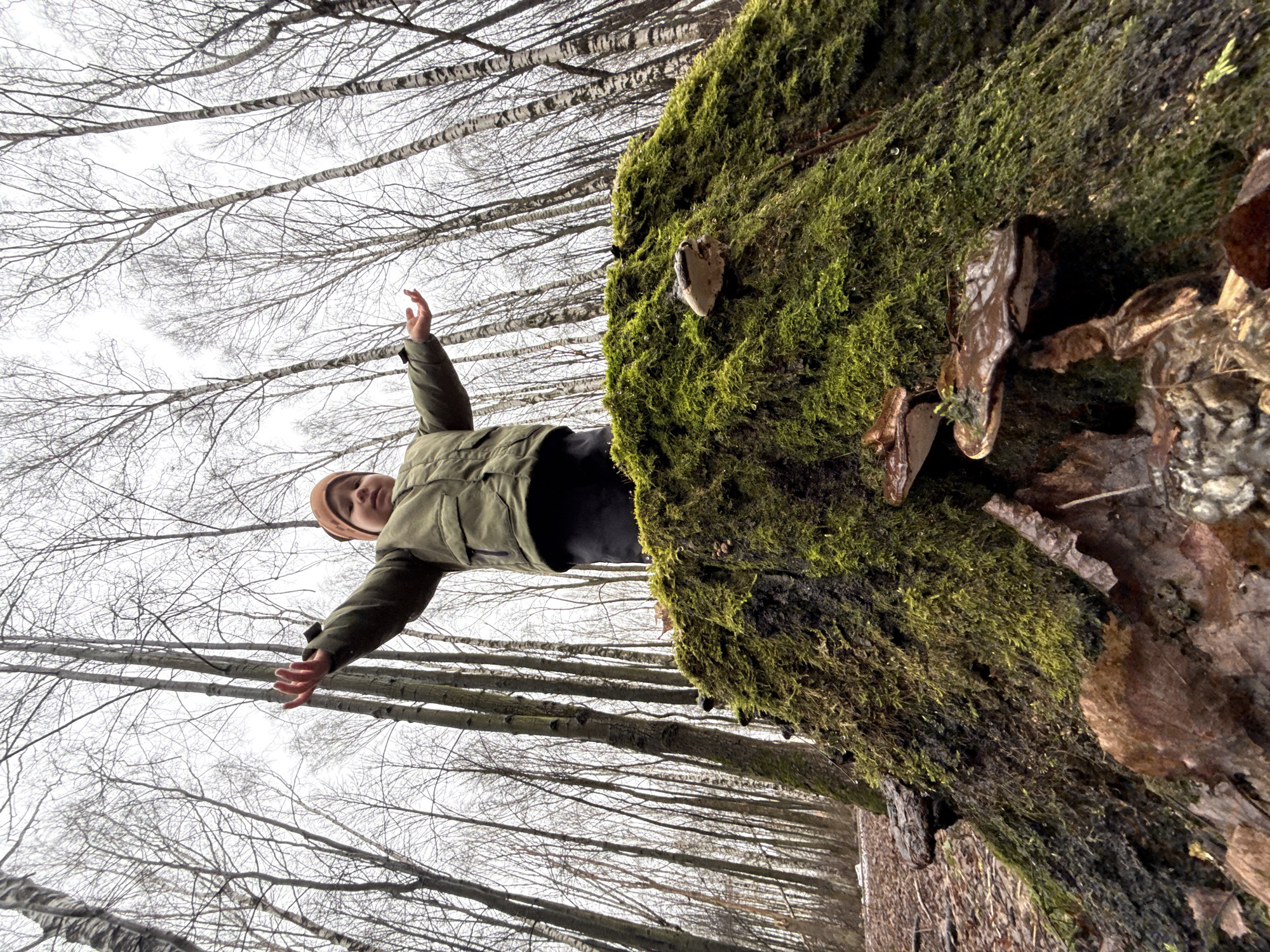 A person stands with arms outstretched near a tree trunk covered in green moss and fungi. The setting is a forest with bare trees visible in the background. The location is Unknown, Unknown.