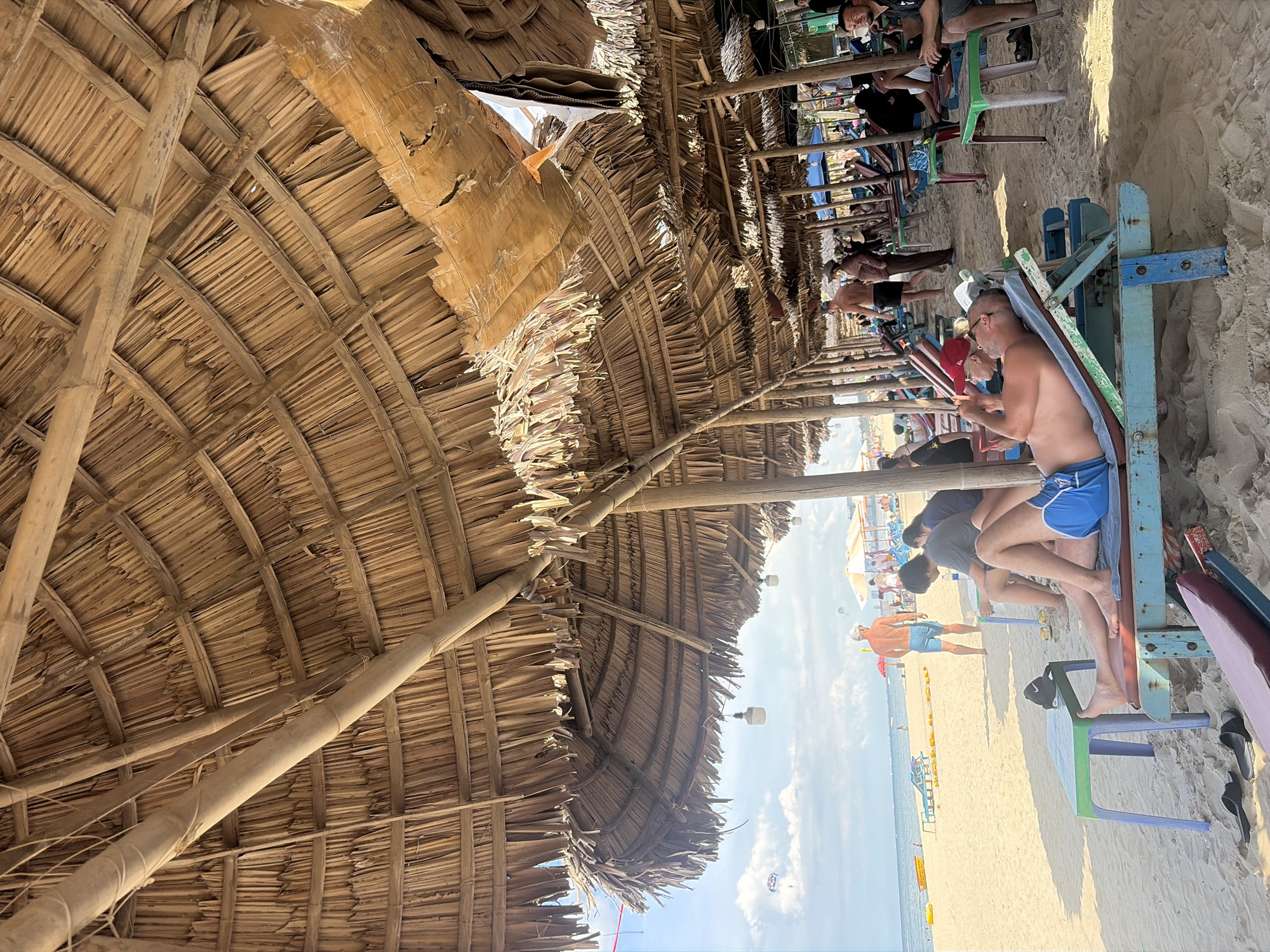 The image depicts a beachfront scene in Da Nang, Vietnam, featuring a row of thatched-roof umbrellas providing shade over beach chairs and tables on a sandy beach. In the foreground, a man is reclining on a blue and green beach chair, wearing blue shorts and holding a phone. To his left, a small green table holds a black baseball cap. Several other individuals are visible in the background, some sitting on chairs or walking along the beach. The ocean and sky are visible beyond the beach umbrellas. The umbrella structure is made of bamboo poles and thatched palm leaves. In the distance, there are colorful beach chairs and a parasailer in the sky. The lighting suggests it is daytime, with sunlight filtering through the thatched roofs.