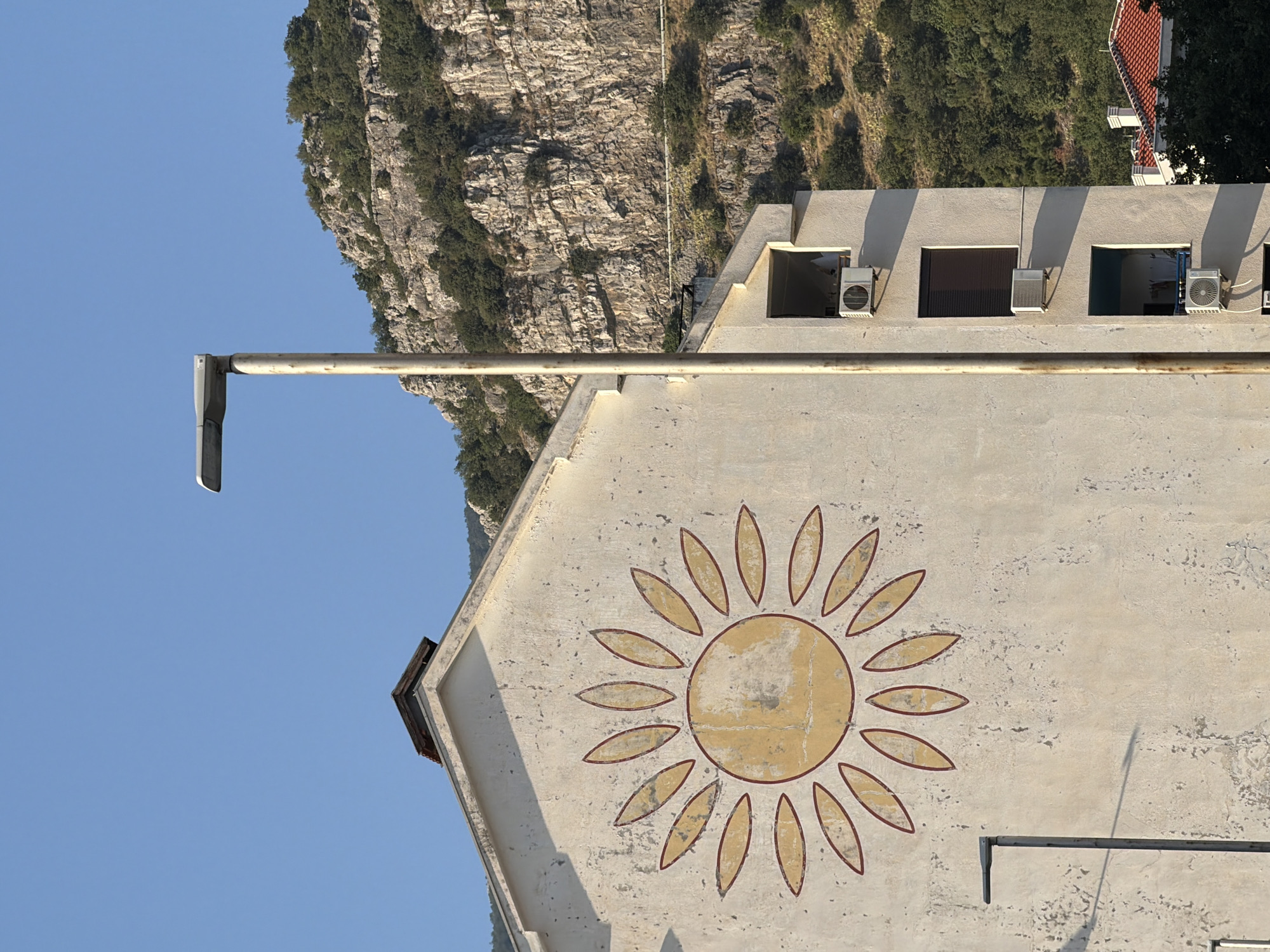 A sun mural adorns the side of a building in Budva, Montenegro, with a rocky, tree-covered mountain forming the backdrop. A tall, gray lamppost stands in front of the building, casting a shadow on the sun mural. The building itself is a pale, textured color, with a triangular roofline at the top. There are several windows visible on the side of the building, some with air conditioning units attached. The sky is a clear, bright blue. The overall scene suggests a sunny day in a Mediterranean coastal town.