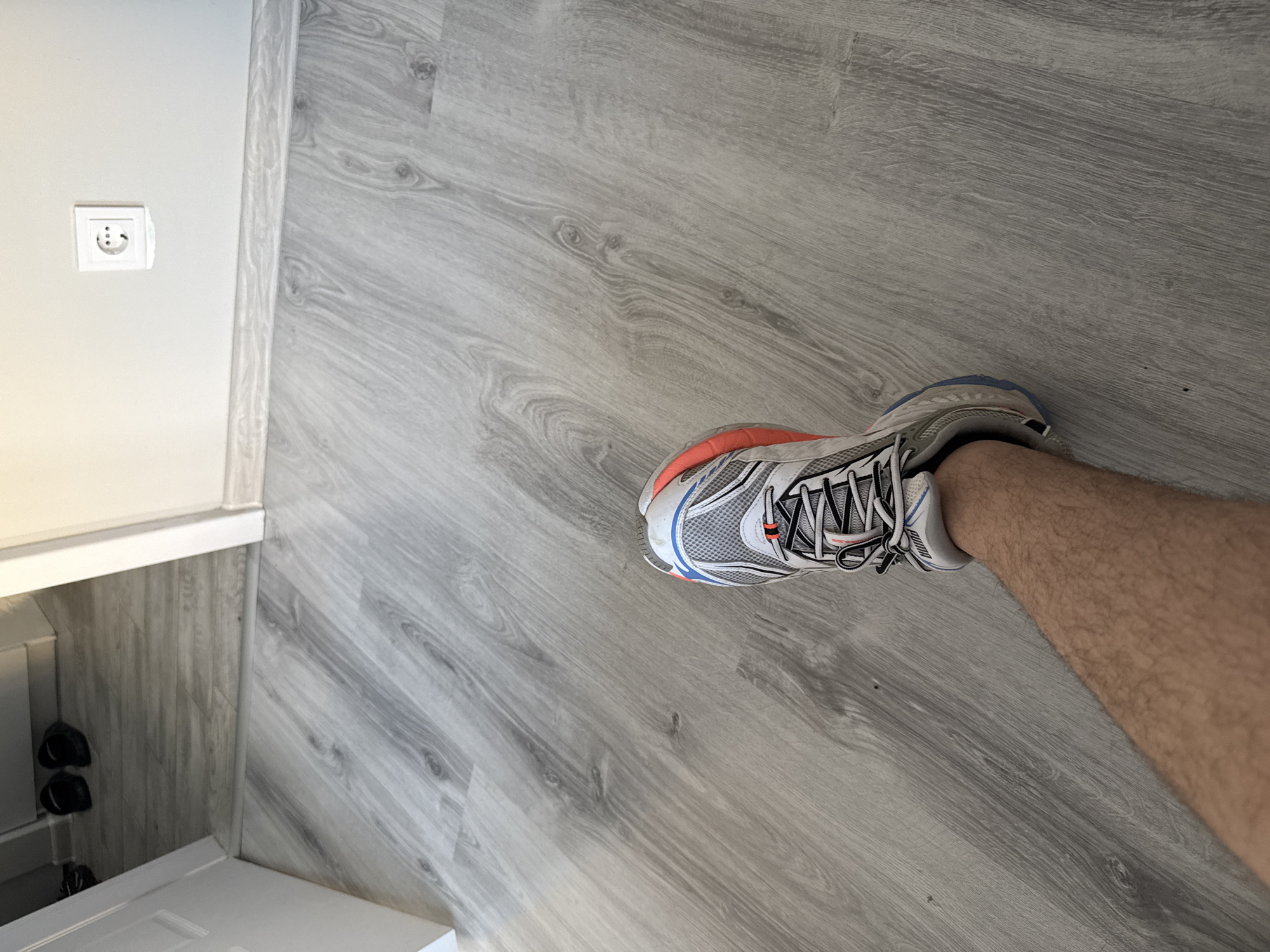 A leg wearing a running shoe is visible against a gray wood-patterned floor. In the background is a white wall with a European-style electrical outlet. The scene appears to be indoors, likely in a room within a building. There is no specific indication of the location being in the city of Stara Zagora, Bulgaria.