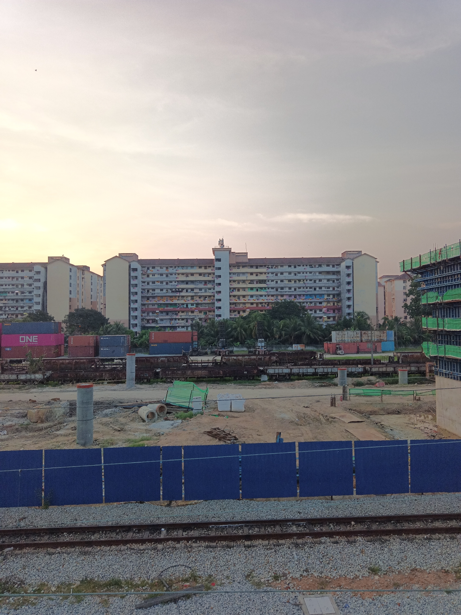 The image depicts a view of apartment buildings and a construction site in Klang City, Malaysia, during a soft light, likely dawn or dusk. In the background, several multi-story apartment blocks, painted in various colors, are visible. In front of the apartments, there are stacks of shipping containers, some with visible branding like "ONE" and "SEALCO." Below the containers, a series of old train carriages are parked on railway tracks. The foreground shows a construction zone with dirt, concrete pipes, a large green mesh structure, and construction materials. A blue barrier fence runs across the middle ground, separating the construction area from the foreground railway tracks. The sky above is overcast with a gentle gradient of light. There are no people visible in the scene.