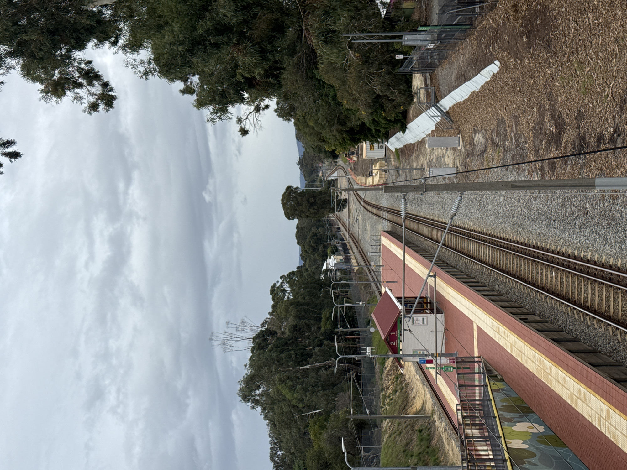 An elevated, downward-angled view shows the Bassendean train station in Bassendean, Australia. The station platform, predominantly in shades of red and tan brick, is visible to the lower right, with a shelter-like structure bearing the number "21" and signage. Tracks run parallel to the platform, extending into the distance, flanked by gravel and foliage. Trees line both sides of the tracks, with a backdrop of hills. The sky is overcast, with heavy cloud cover. Overhead electrical lines and support structures are visible along the tracks. A set of stairs leads down from the platform, and there is a decorative design on the lower portion of the platform.