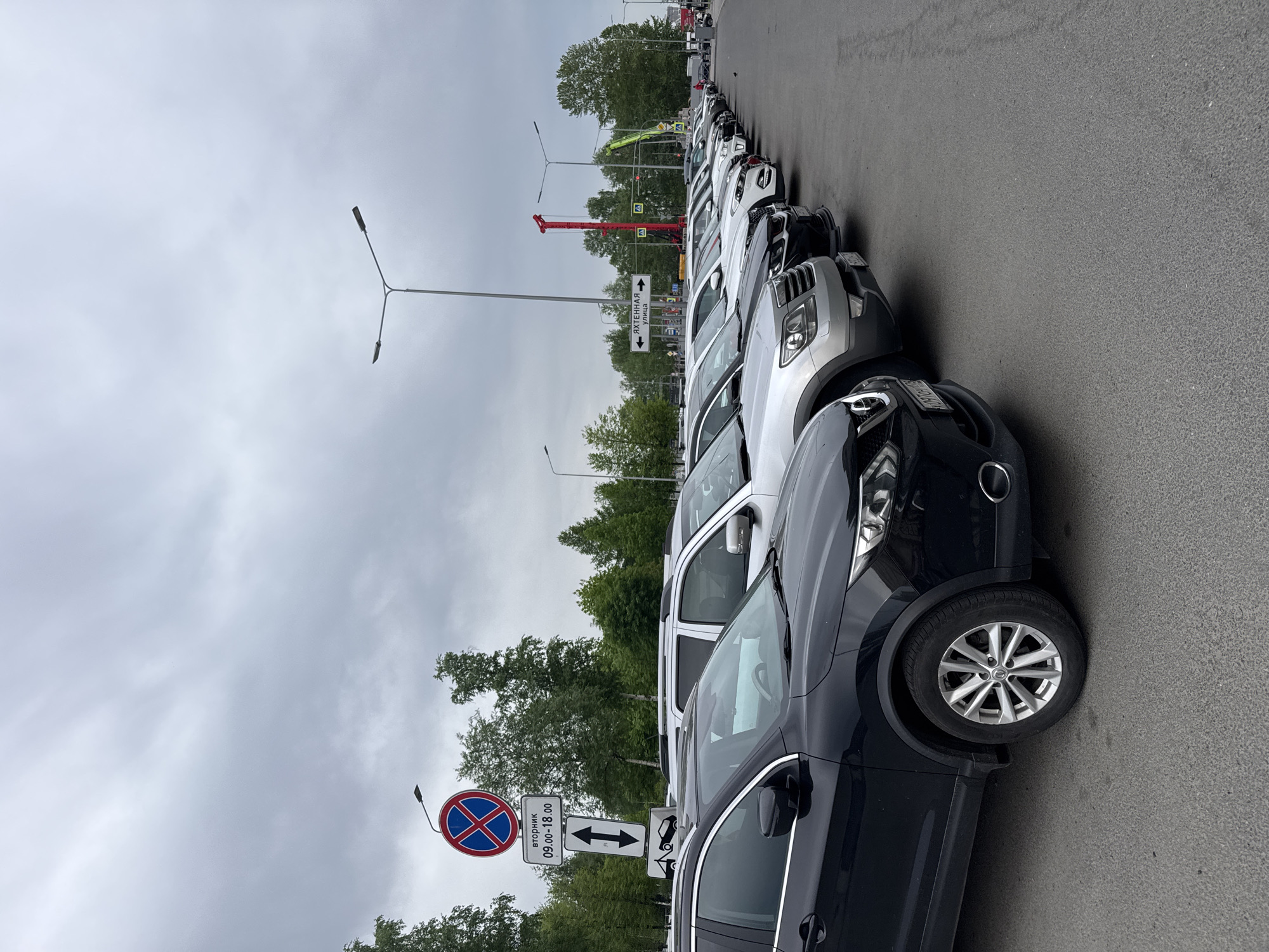 The image depicts a row of parked cars along a street in what appears to be Saint Petersburg, Russia. The cars are various models and colors, predominantly white, silver, and black. A street sign in Russian indicates parking restrictions between 9:00 and 18:00. Another street sign points to "Яхтенная улица". A red crane is visible in the background. The sky is overcast. There are several trees in the background.