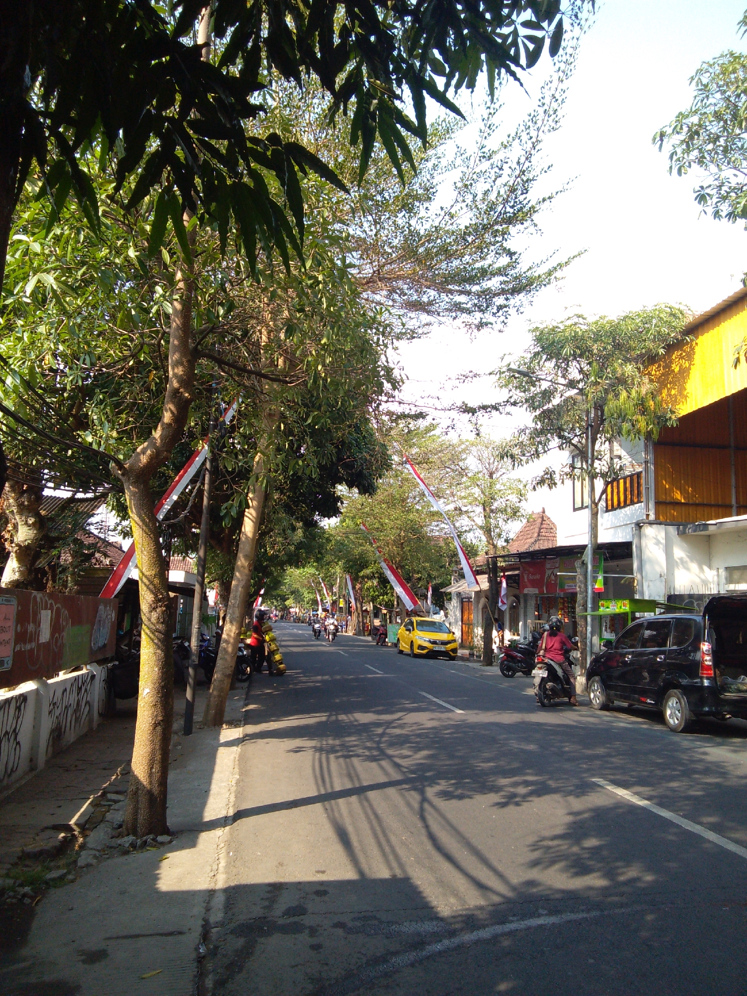 This is a street scene in Trenggalek, Indonesia, during daylight. The image captures a road lined with trees and buildings on both sides. Several vehicles are visible, including a yellow car, a black SUV, and multiple motorcycles. Some people are on motorcycles, and others are near the buildings. Red and white flags are displayed along the street, possibly indicating a local celebration or event. The environment appears to be a typical urban setting with some greenery.