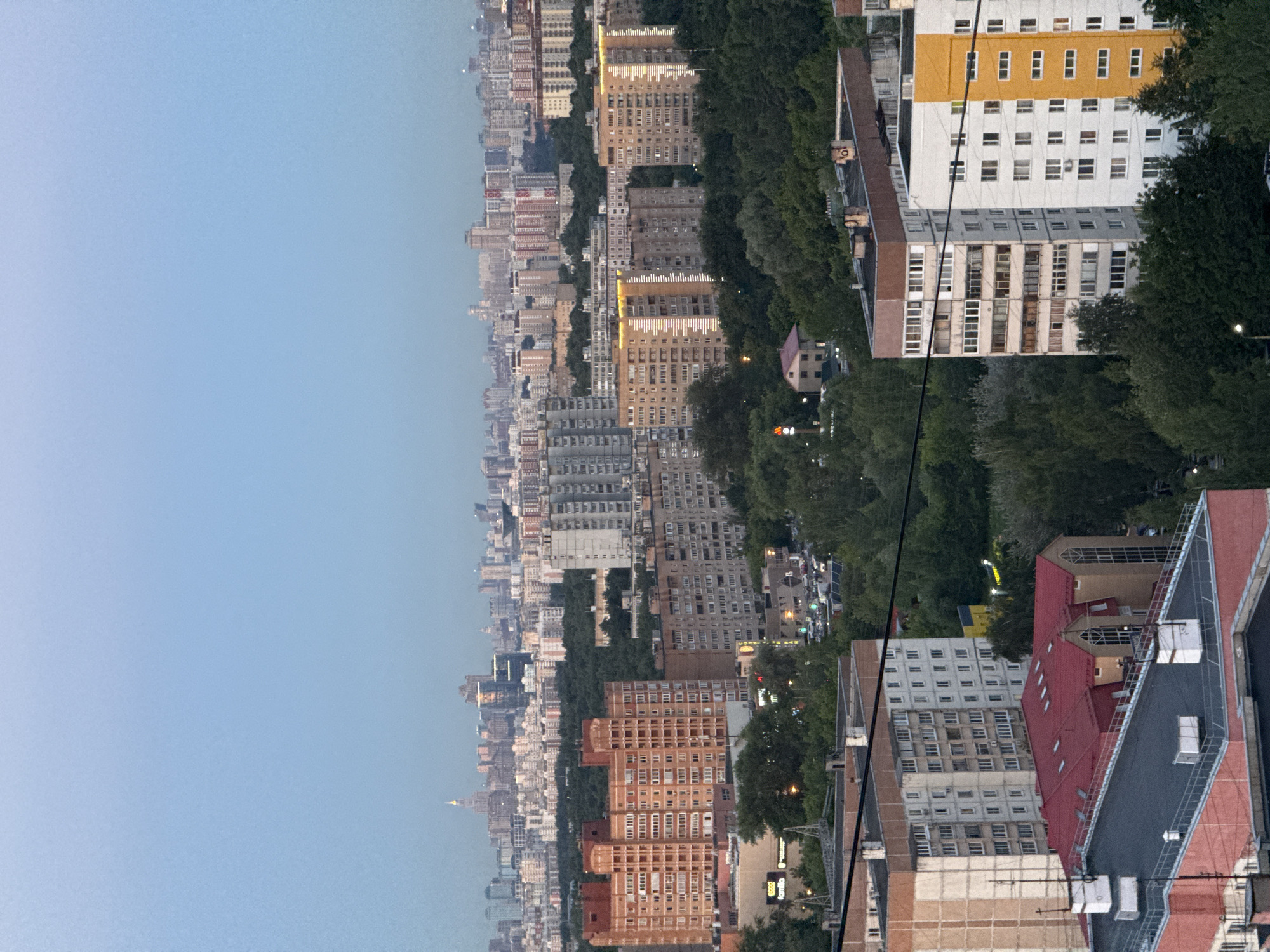 This is a vertical, medium-range, eye-level shot of a cityscape in Khimki, Russia. The image captures a dense urban environment with a variety of buildings, ranging from older, utilitarian structures to more modern apartment complexes. Trees are interspersed throughout the scene, creating a contrast between the natural and man-made elements. The sky is a light, hazy blue, suggesting it may be early morning or late evening, or perhaps overcast.
In the foreground, the roofs of several buildings are visible, some with red metal roofing and others with darker materials. One building in particular has a striking orange and white facade with many regularly spaced windows. Further back, a long line of buildings stretches into the distance, with varying architectural styles and heights. Some of these buildings have illuminated windows, indicating activity within. The density of the buildings suggests a populated urban area.
A prominent feature is a cluster of tall, slender trees that line the side of a street or park area, separating some of the buildings. The overall impression is one of a bustling city, possibly at dusk or dawn, with the sky suggesting a calm or perhaps slightly humid atmosphere. There are no people clearly visible in the frame, and the focus is on the architecture and the urban landscape. The image has a slightly desaturated quality, which could be due to the lighting conditions or post-processing.