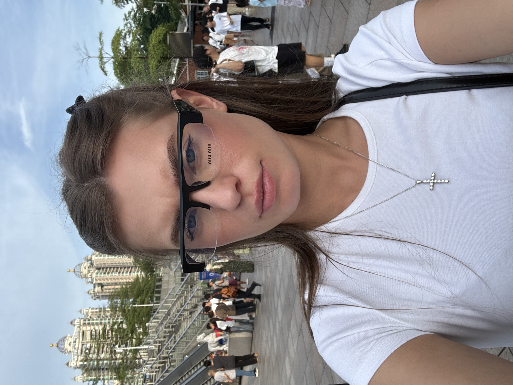 An outdoor selfie taken by a young woman with light brown hair and blue eyes wearing black-rimmed glasses and a white t-shirt with a silver cross necklace. She is in Haizhu District, China. In the background, there are several people walking around, and a modern building with domes can be seen. The sky is cloudy.
