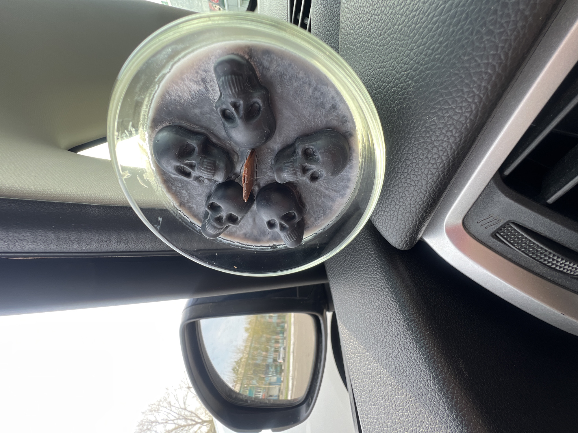 A close-up interior shot focuses on a candle containing multiple black skull figurines embedded in grey wax, sitting in a clear glass jar. The candle is situated inside a car, next to an air vent with a silver trim and black slats. The car's side mirror is visible below, reflecting buildings and trees, indicating an urban or suburban environment. The interior surfaces of the car surrounding the candle are primarily black with a textured finish. The environment outside is bright, suggesting daytime.