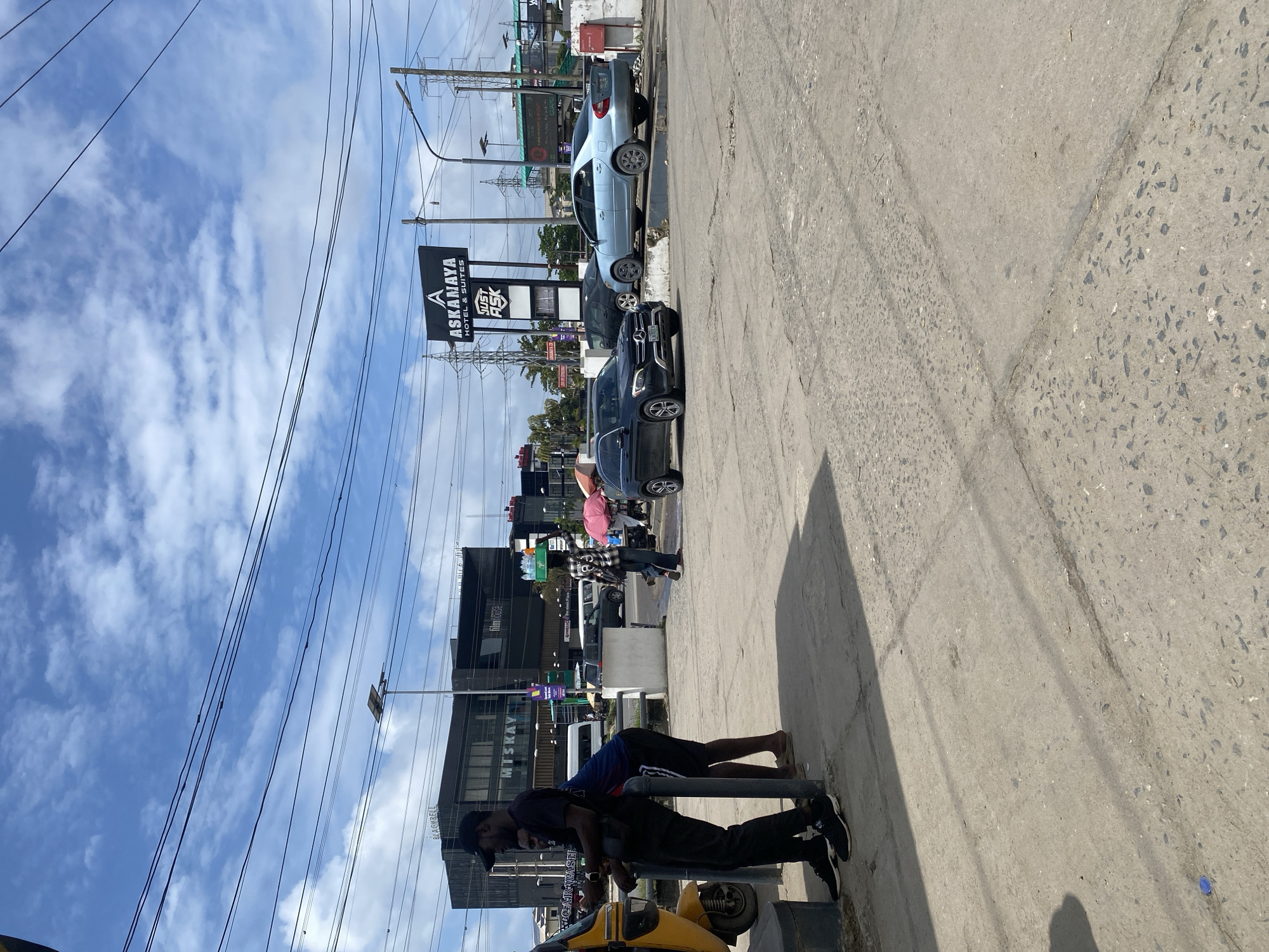 In Ajah, Nigeria, a street scene unfolds under a partly cloudy sky. A yellow tricycle sits parked on the left. Two men stand near the tricycle, one in a black t-shirt and black pants, wearing a black cap, and the other in a blue t-shirt and black shorts. In the background, a modern black building with "MISKAY" and "FILMHOUSE" signs is visible. Further down the street, a prominent sign for "ASKANMAYA HOTEL & SUITES" with "JUST ASK" below it stands tall. Several cars, including a dark Mercedes-Benz SUV and a light blue sedan, are present on the road. Other vehicles, including vans and a white minibus, are also visible. A person is seen walking with a tray of bottled drinks balanced on their head. The environment is characterized by paved roads, concrete sidewalks, overhead power lines, and streetlights.