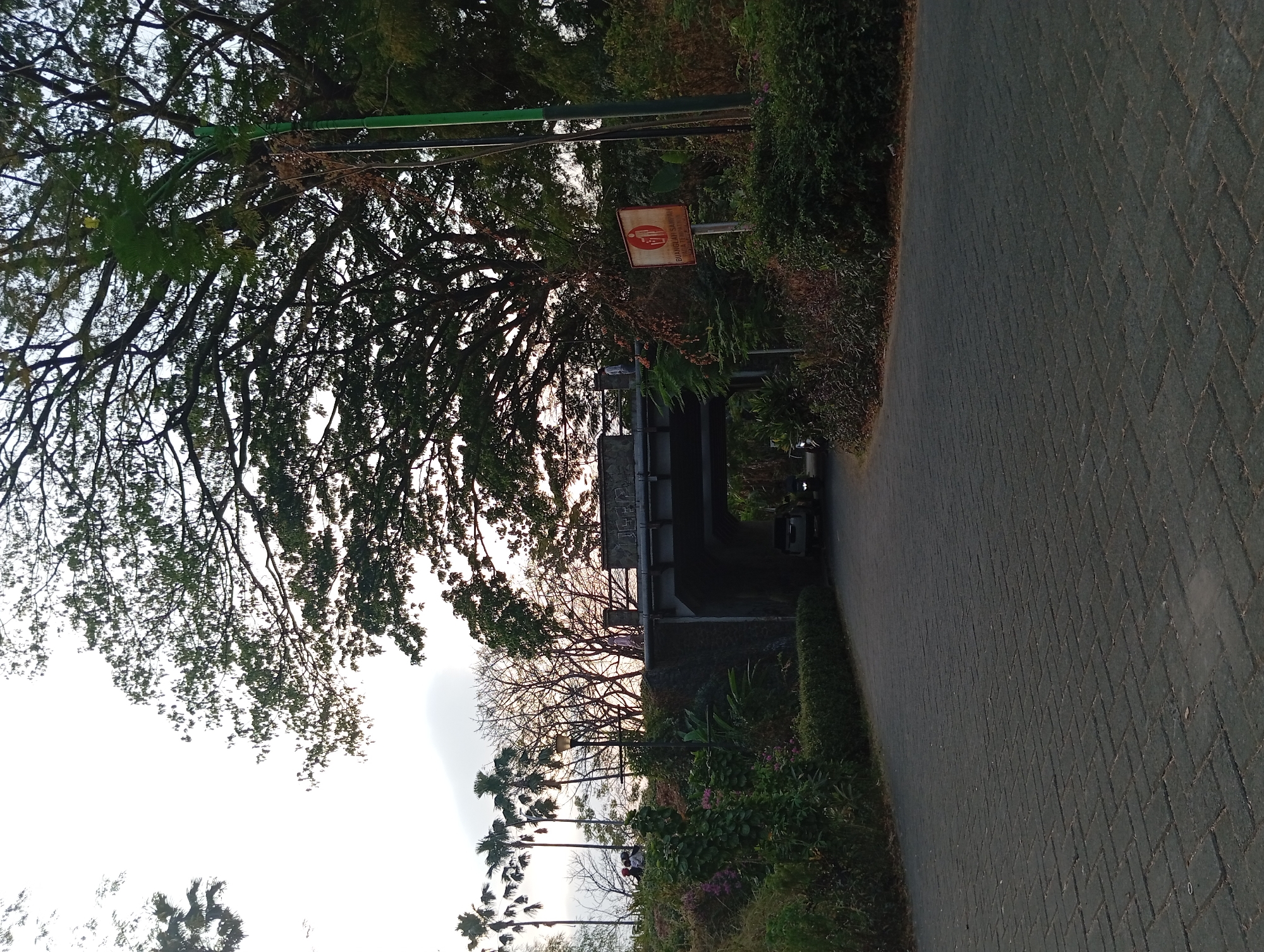 The image shows a paved road in the city of Karang Jati, Indonesia, leading towards an underpass. The road is made of dark gray interlocking bricks, and on either side, there are green bushes and trees. A large, mature tree with sprawling branches dominates the upper right portion of the image. Above the underpass, there is a bridge with a sign that appears to have graffiti on it. On the bridge, a few people are visible, and one person is walking. To the right of the underpass, a red and white sign is posted on a pole, depicting a person throwing trash into a bin, with Indonesian text below. Inside the underpass, a white SUV and a motorcycle are seen. The sky is overcast with some sunlight filtering through. The overall scene suggests a public area, possibly a park or a road within a residential or recreational zone.