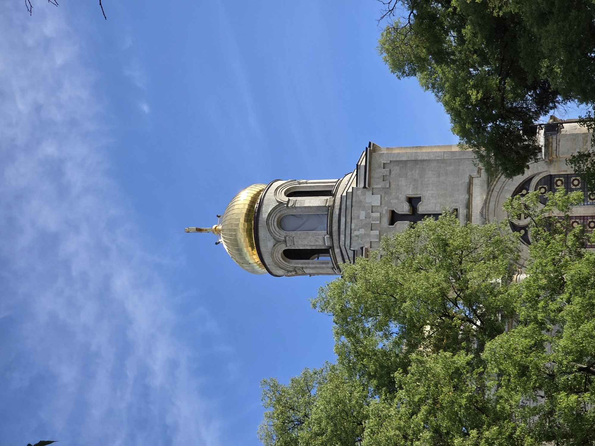 The image shows the bell tower of the Dormition of the Mother of God Cathedral in Varna, Bulgaria. The golden dome of the tower is visible against a blue sky. Green trees surround the base of the tower. The tower is made of light-colored stone and features arches, a cross, and other architectural details.
