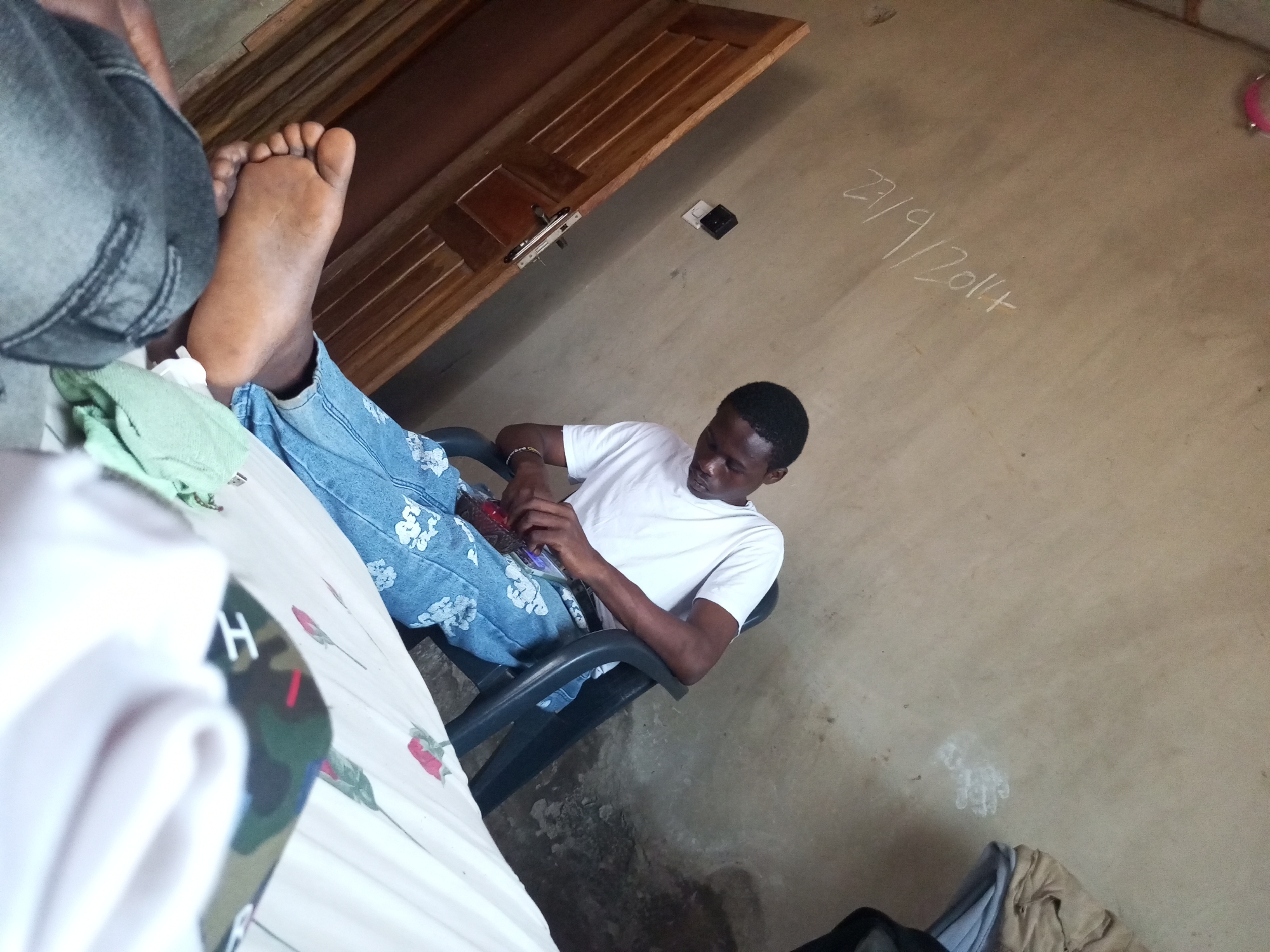 In an indoor environment, a Black male is seated in a black chair, wearing a white t-shirt and blue jeans. He is holding a red object in his hands and appears to be focused on it. To his left, a person's legs are visible, with one bare foot resting on a white surface. A wooden door is seen in the background, and a date written in chalk, "27/9/2014", is present on the wall. The floor is concrete, and there is some debris scattered around. The overall lighting suggests an indoor setting with artificial light.