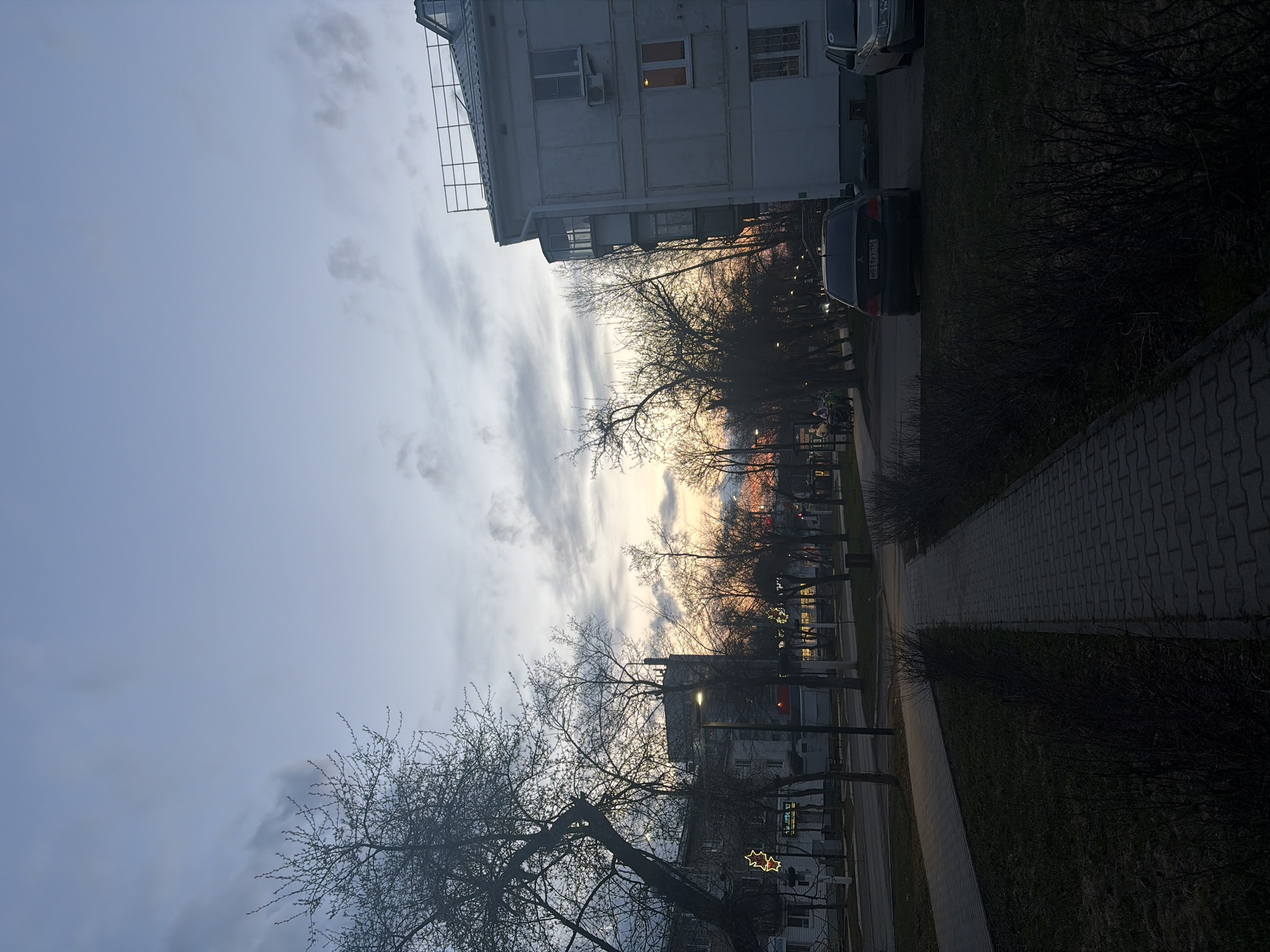 The image captures a street scene at dusk, likely in Chaikovsky, Russia. The sky dominates the upper left, displaying a soft blue transitioning into warm orange and pink hues of a sunset near the horizon, partially obscured by bare tree branches. Wispy clouds are scattered across the sky.
On the right side, a multi-story residential building with a white facade and visible windows, some illuminated, stands against the fading light. An external air conditioning unit is visible on its wall. Below the building, several leafless trees with intricate branch patterns line what appears to be a street or pathway. Further along, more trees, streetlights, and smaller buildings are visible in the distance.
A paved sidewalk with a distinctive brick pattern runs along the bottom right, alongside a strip of grass and dark bushes. Parked cars are visible, including a dark sedan with a clear license plate, "M911YY 196 RUS," which indicates a Russian registration from the Sverdlovsk Oblast region.
The scene depicts a tranquil evening, with artificial lights beginning to glow, including a distinct red star-shaped decorative light mounted on a pole near a building in the middle ground. The bare trees suggest either late autumn, winter, or early spring. No significant activity is explicitly captured beyond the general ambiance of an urban residential area winding down for the night.