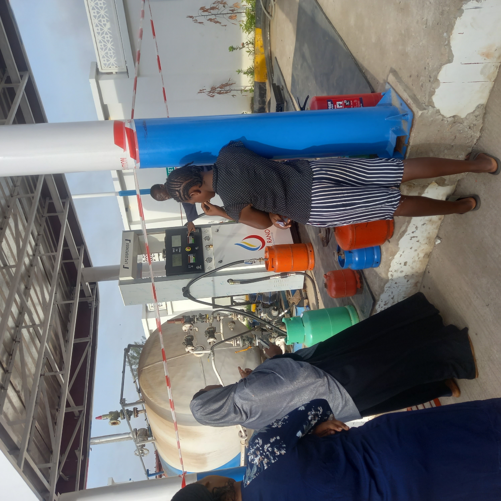 The image shows a gas station in Barden Yana, Nigeria. A young woman in a black and white striped skirt and a polka dot top stands near an LPG dispenser, possibly operating it. Several colorful LPG tanks (orange, blue, green) are scattered nearby. Two women in hijabs are visible in the foreground, observing a large, cylindrical tank with pipes and valves. A red fire extinguisher is also present. The gas station has a metal frame structure and is surrounded by a white wall. The sky is visible in the background.