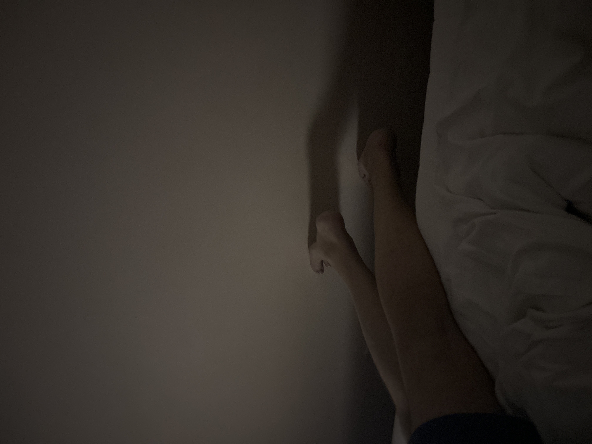 The image captures a dimly lit indoor scene, likely a bedroom, with a focus on a person's legs and feet. The setting appears to be nighttime or a very dark room, indicated by the low light and deep shadows.
On the right side of the frame, a person's legs and feet are visible. The skin tone is light to medium. One foot is distinctly seen bent against a wall or headboard, and the toenails appear to have a dark polish. Below the knees, a portion of dark blue fabric, possibly shorts or underwear, is visible. The person's lower body is partially covered by a white, rumpled blanket or duvet, which occupies the lower right and center-right of the image. The folds and texture of the bedding are subtly visible despite the darkness.
The left two-thirds of the image consist of a plain, dark gray wall, which shows some subtle variations in shading, possibly from ambient light sources or shadows cast by objects outside the frame. The wall serves as a stark, minimalist background, emphasizing the figure in the bed.
The activity taking place suggests the person is resting, possibly sleeping, in bed. There are no other objects or specific details that provide additional context about the immediate surroundings within the room. No text is visible in the image. The location context provided is Budva, Montenegro.