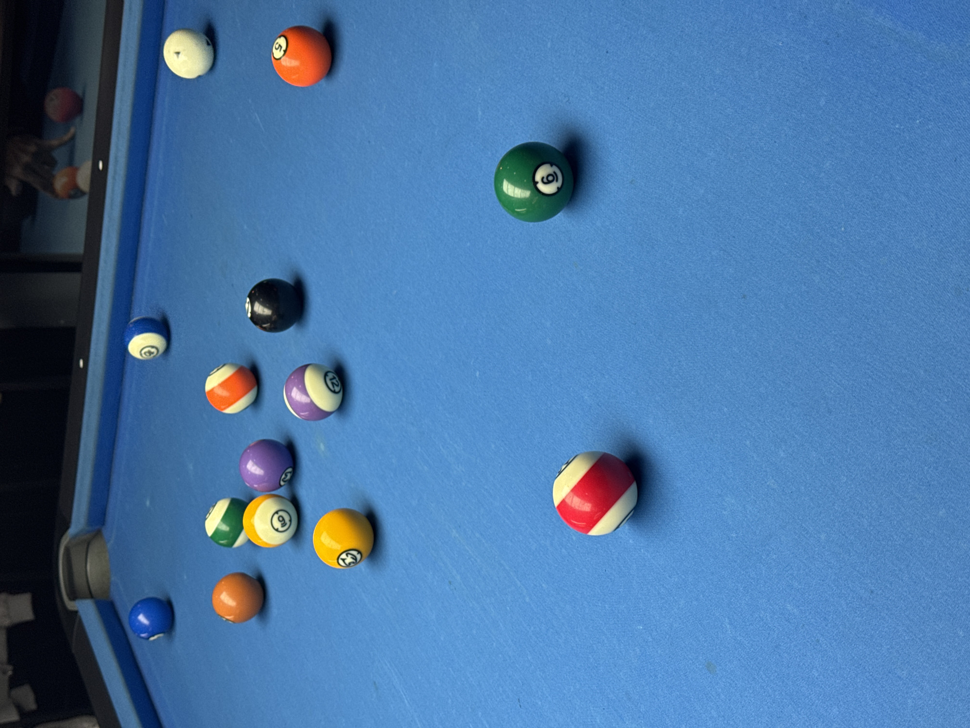 The image depicts a billiard table with multiple pool balls scattered across its blue surface. The balls are numbered and colored. The table's rail is visible along the left edge, and part of the room in which the table is located can be seen in the background. There is nothing present in the image that could be used to determine its location. The image cannot be determined to be taken in the city of Bellville, South Africa.