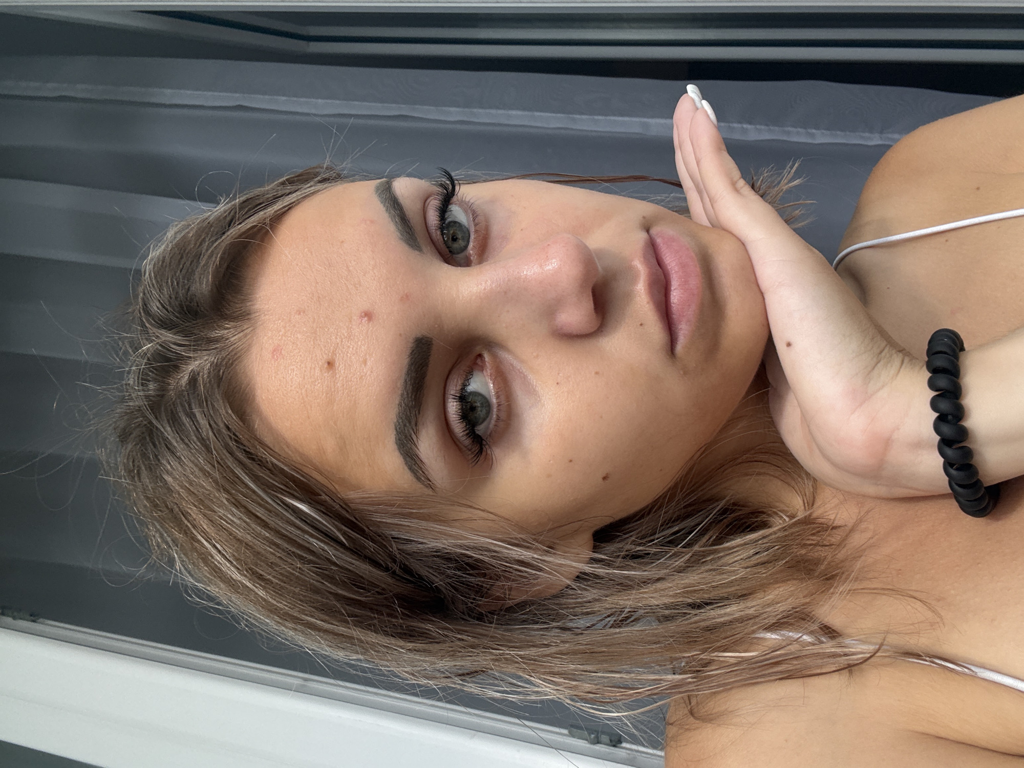 A young woman with light brown hair is resting her chin on her hand. Her hand is positioned under her chin and cheek, with her fingers spread out. She is wearing a white strap top. Her hair is damp and messy, falling around her shoulders. Her eyes are looking directly at the camera, and she has prominent eyelashes. There is a slight sheen on her skin, and a few blemishes are visible on her forehead. The background appears to be indoors, with vertical blinds or curtains behind her. There is no indication of the city of Kholmsk, Russia, in the image.