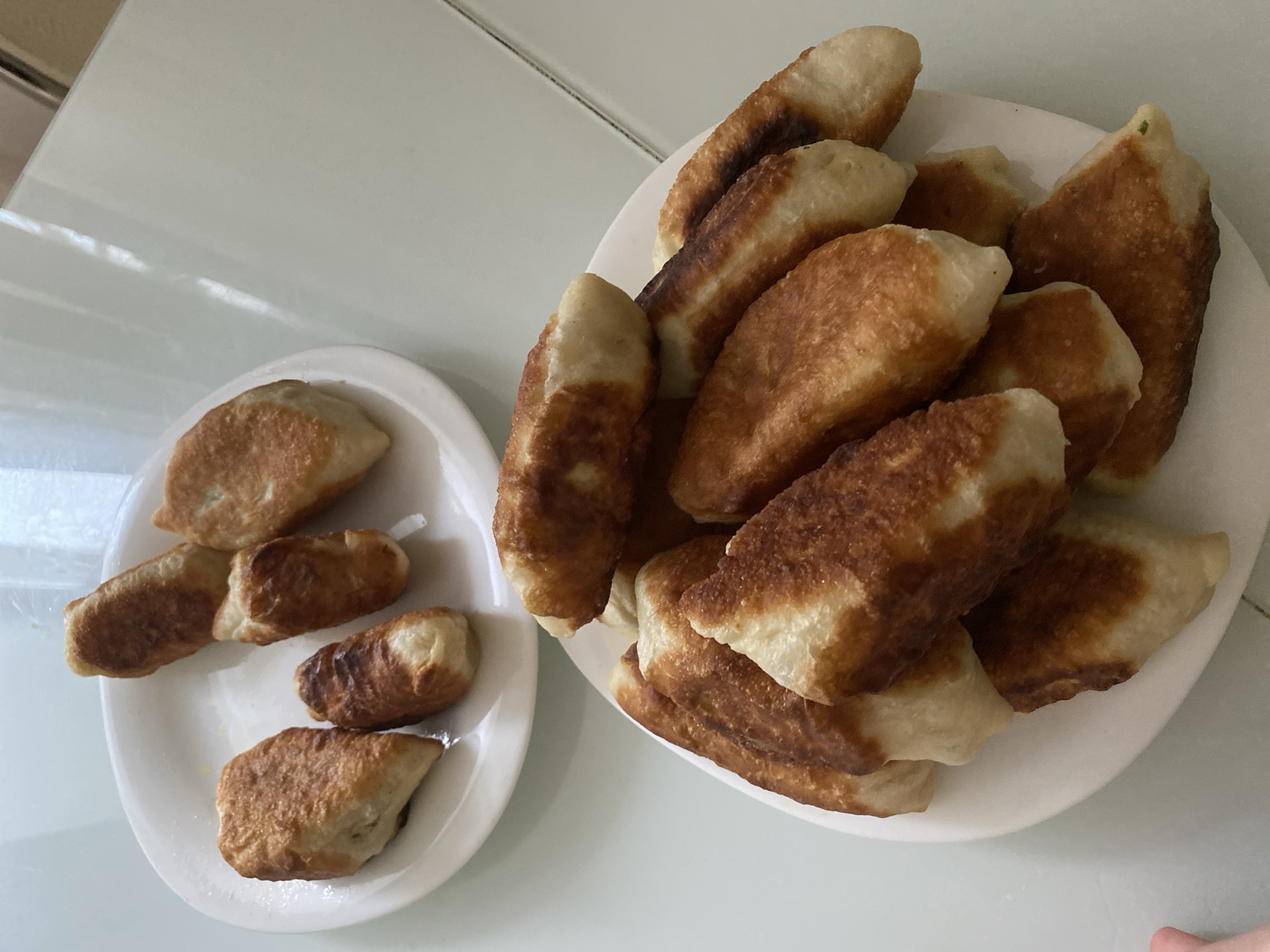 The image shows two white plates of fried dumplings on a white surface, likely a table or countertop. The larger plate on the right is piled high with approximately 15-20 dumplings, while the smaller plate on the left has about 5 dumplings arranged more sparsely. The dumplings are golden brown, suggesting they have been fried. The image does not provide any information about the city of Votkinsk, Russia, people, actions or interactions.