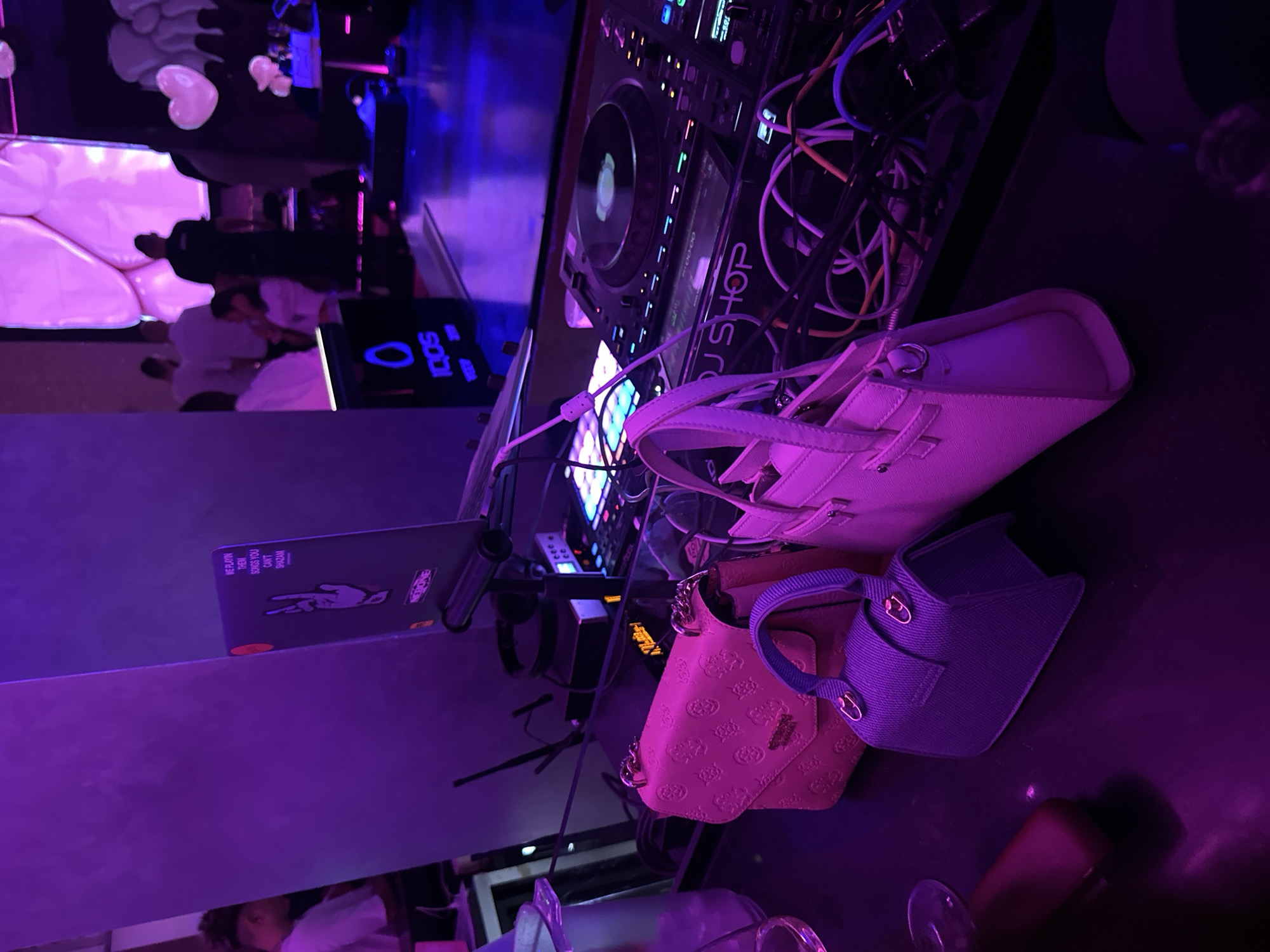 The image shows a nightclub scene, likely in Sofia, Bulgaria, judging from the "IQOS" logo and the text "SHOP" visible on the DJ equipment.
The scene focuses on a DJ setup, including a DJ mixer, laptop, and associated cables. The laptop displays a sticker with the phrases "WE PLAYIN THEM SONGS YOU CANT SHAZAM" and "NEBONE."
Scattered near the equipment are three handbags: one light-pink, one light-peach with a Guess logo, and one blue.
In the background, there is a blurry view of other club patrons and lighting, suggesting a social gathering in a nightclub setting.