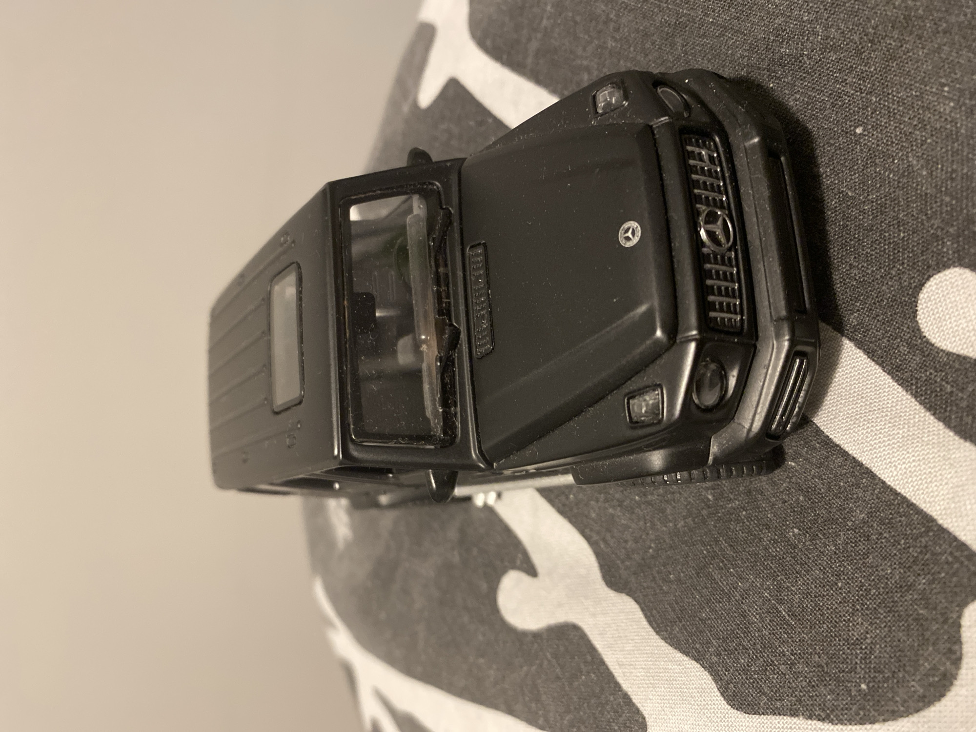 A black toy Mercedes-Benz G-Wagon is displayed on a patterned surface. The toy car is black with silver accents on the grille and emblem. The background is a plain, light-colored wall. There is no indication of the city of Łódź, Poland, or any people present in this image.