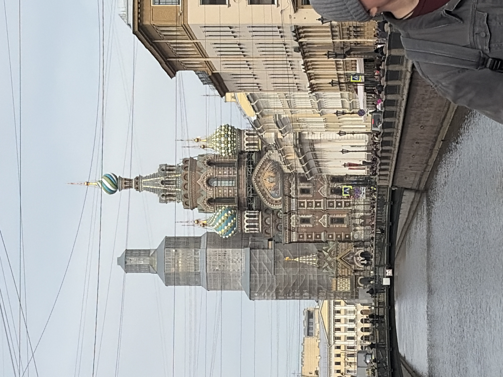 The image captures the Church of the Savior on Spilled Blood in Saint Petersburg, Russia, as seen from a bridge over a canal. The church's distinctive, multi-colored onion domes and ornate facade are prominent. Scaffolding partially obscures a tower of the church. A canal runs along the right side of the image. People can be seen walking along the bridge and the embankment. The sky is overcast. Power lines are visible in the foreground. A person wearing a gray beanie and jacket is partially visible in the upper right corner of the frame. Other buildings are also visible in the background.