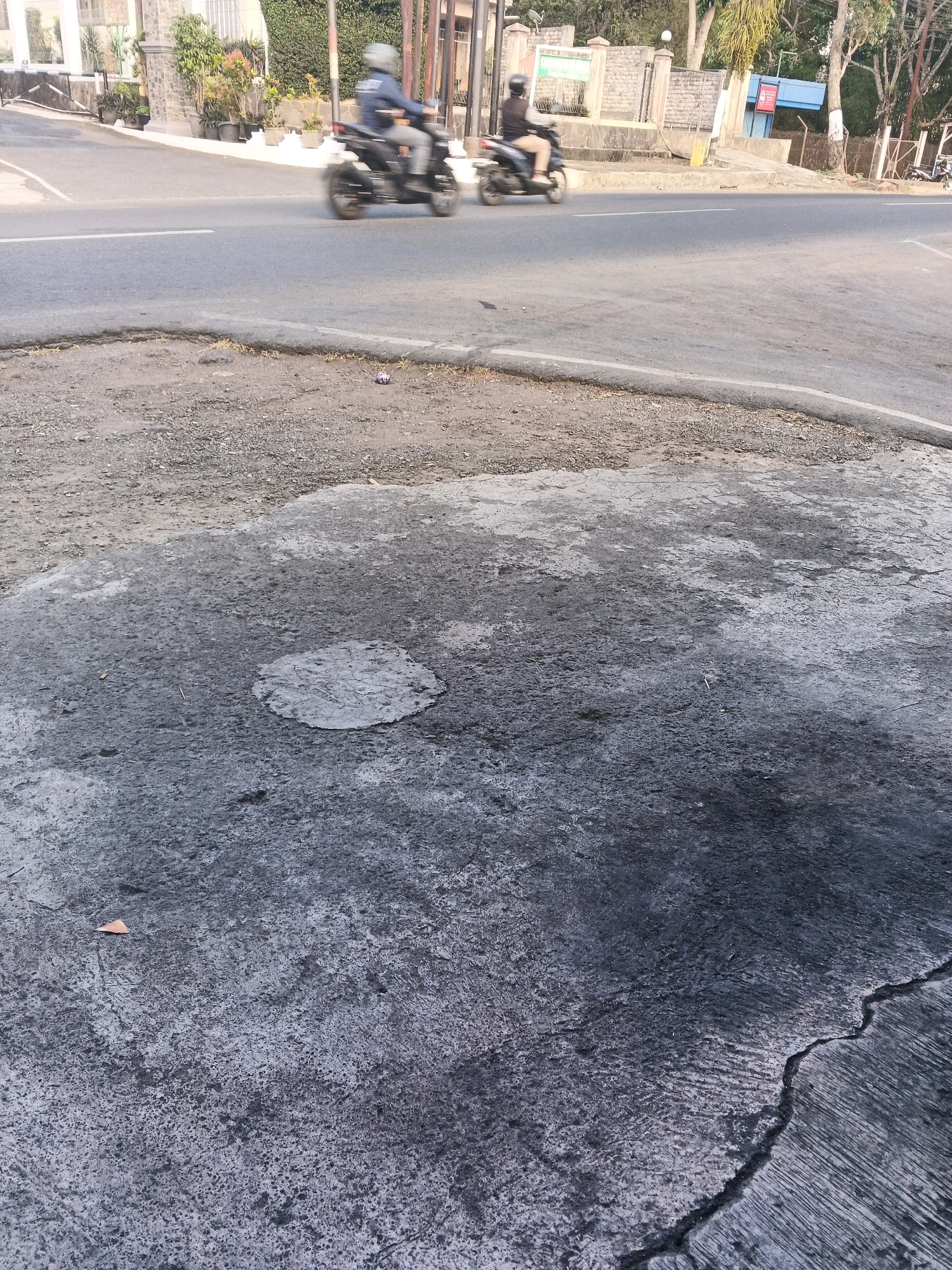 The image depicts a street scene with two individuals on motorcycles blurred due to motion, traveling on a paved road. The foreground features a rough, textured asphalt surface with patches of lighter grey and darker grey, indicative of wear and tear, including a circular indentation filled with lighter material and several cracks. The background shows a sidewalk area with potted plants and buildings, including a structure with a blue facade and a fence. The context of Semarang, Indonesia, suggests a tropical environment, though this is not explicitly visible beyond the vegetation.