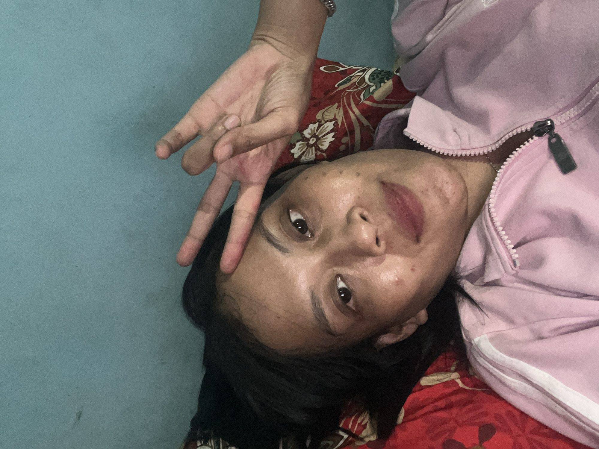 The image shows a selfie of a woman lying down and making a peace sign with her right hand. She is wearing a light pink zip-up jacket. The background consists of a teal-colored wall and a pillow with a red floral pattern. The woman has dark hair and appears to have a light complexion with some skin blemishes. The overall impression is of a casual, indoor setting. The image does not contain any information about the city of Kecamatan Pati, Indonesia.