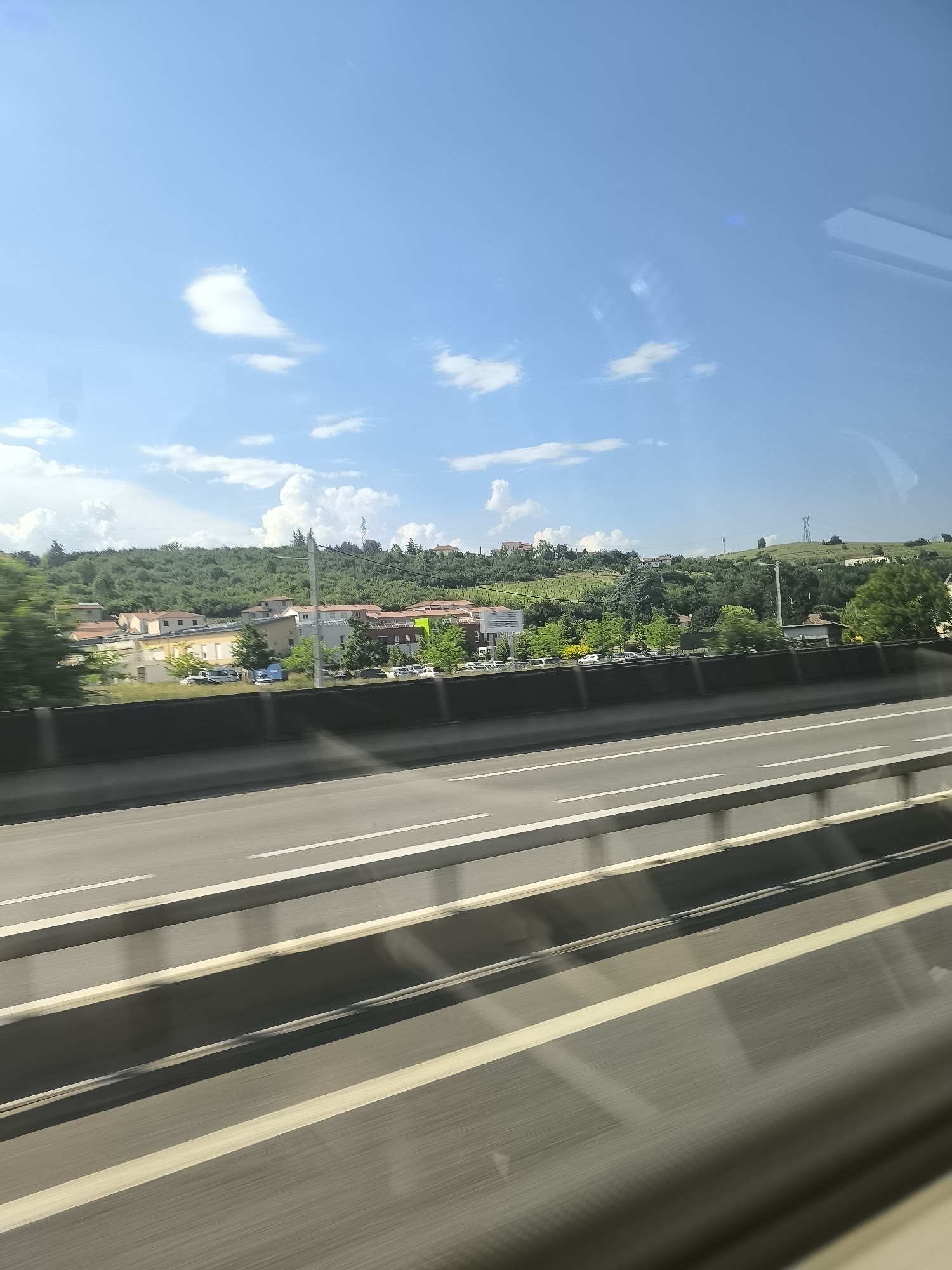 The image, likely taken from a moving vehicle, shows a highway in La Grand-Croix, France. The foreground consists of a multi-lane highway with gray concrete barriers and white lane markings. Beyond the highway is a lower concrete wall, behind which are several buildings with red tile roofs surrounded by trees. Further in the background, a hillside covered in green vegetation is visible under a blue sky with scattered white clouds. There are also some telephone poles on the hill. The overall lighting suggests it is daytime.