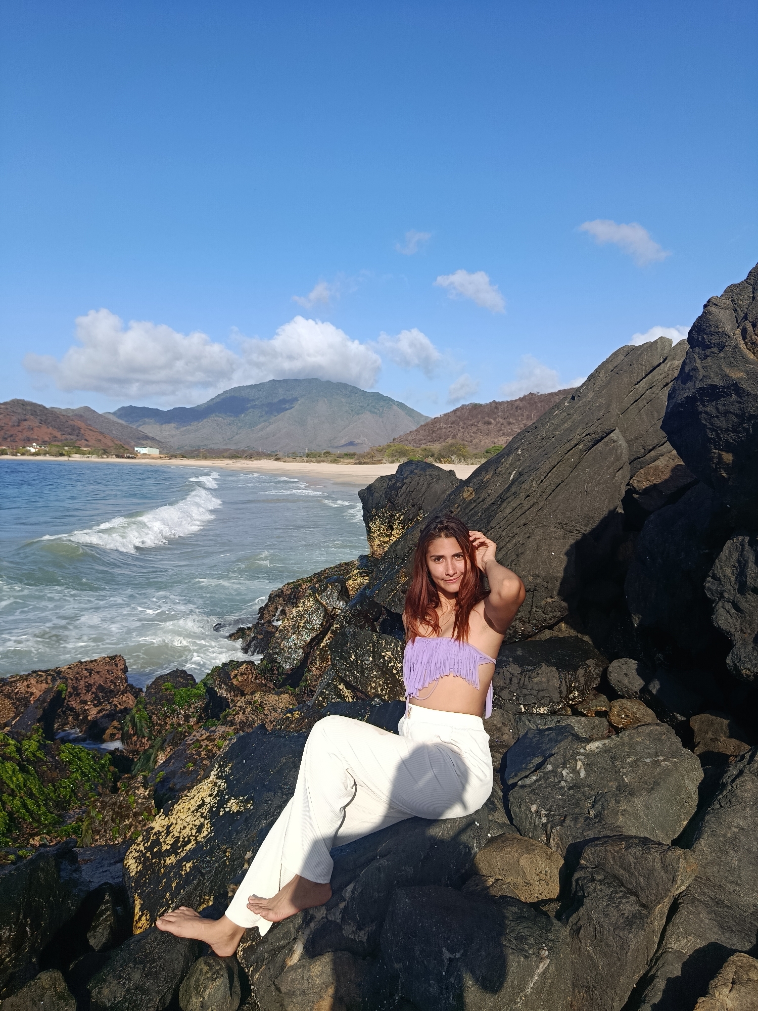 A young woman with long, auburn hair is sitting on dark, jagged rocks on a beach with a turquoise ocean and white waves. She is wearing a light purple, fringe-trimmed bikini top and loose white pants. Her left leg is extended, and her right leg is bent. She is looking at the camera with a slight smile and her right hand is near her hair. The background shows a sandy beach, hills covered in dry vegetation, and mountains under a bright blue sky with scattered white clouds. The rocky foreground is partially covered in patches of green moss and yellow lichen. The scene appears to be in the vicinity of Pedro González, Venezuela.