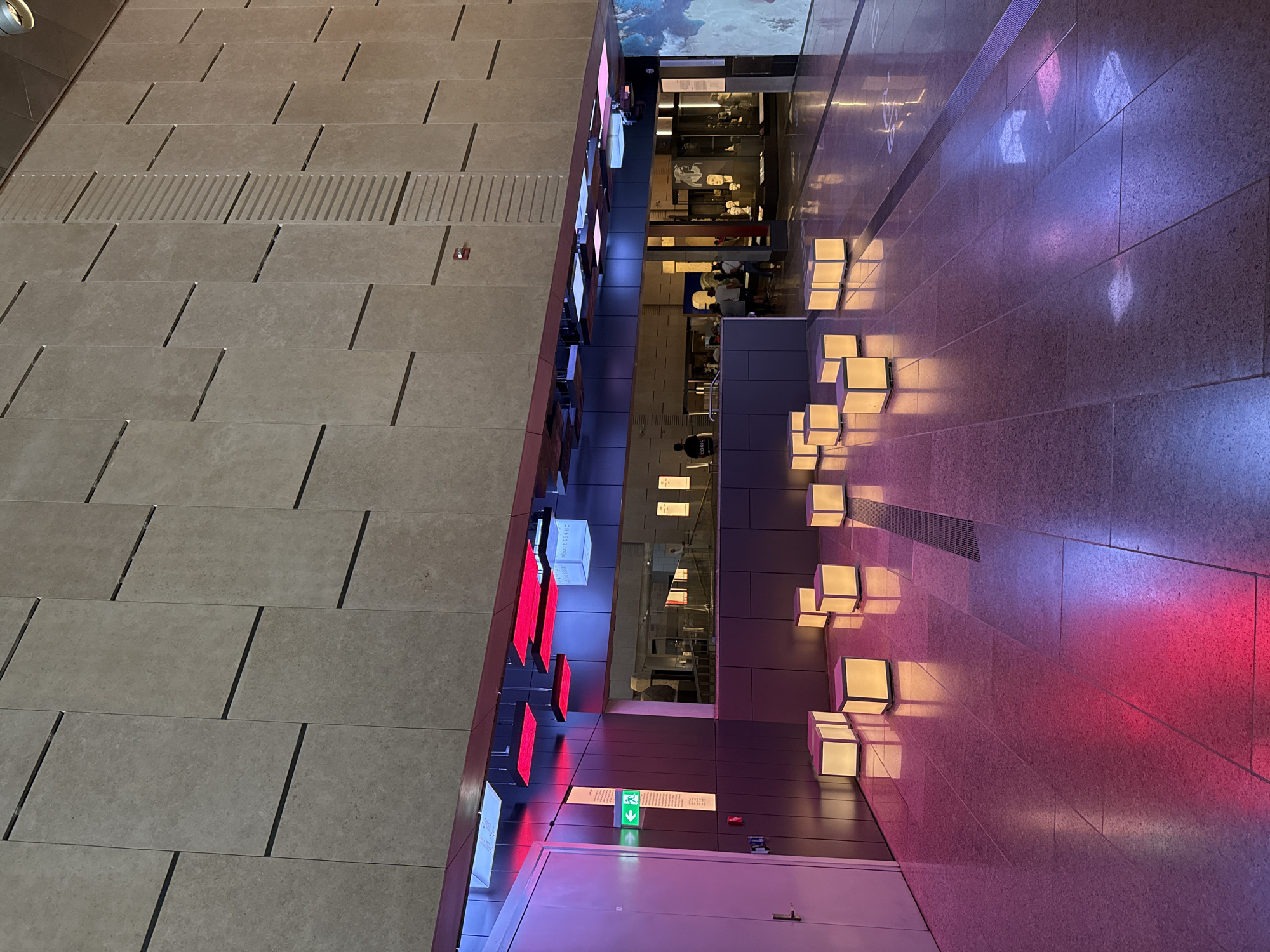 The image depicts an indoor public space, likely a museum or exhibition hall. The architecture features a large, textured stone wall with horizontal dark lines. Below this wall, a darker, reflective surface forms a display area or counter. Suspended above this area are numerous rectangular light fixtures, some emitting bright red light, and some glowing white with text.
In the foreground, several illuminated cubic objects are placed on a polished tiled floor. These cubes emit a soft yellow light and cast reflections on the floor, which is also illuminated with pink and purple hues from the overhead lighting and possibly the cubes themselves.
To the left, a plain white door is visible, with an illuminated green exit sign above it. In the background, through the display area, a crowd of people can be seen, some appearing to be museum visitors looking at exhibits. The overall atmosphere is modern and softly lit, with a focus on the illuminated display elements.
The location specified, Kafrat Nassar, Egypt, is not visually identifiable within the image itself, as the image contains no specific landmarks or textual information to confirm this geographical detail.