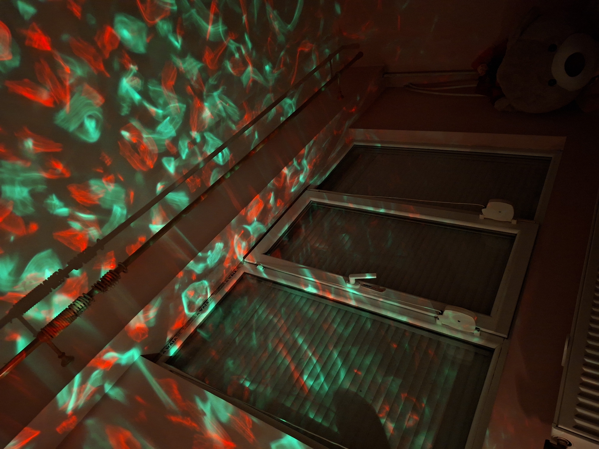 A low-angle, indoor photograph shows a window, a curtain rod, and a stuffed animal. The window is composed of three stacked sections. The window and surrounding wall are illuminated with a pattern of red and green light. A metal curtain rod extends diagonally across the upper-left of the scene. A portion of a stuffed animal is in the upper-right. The location appears to be in a room in Pleven, Bulgaria.